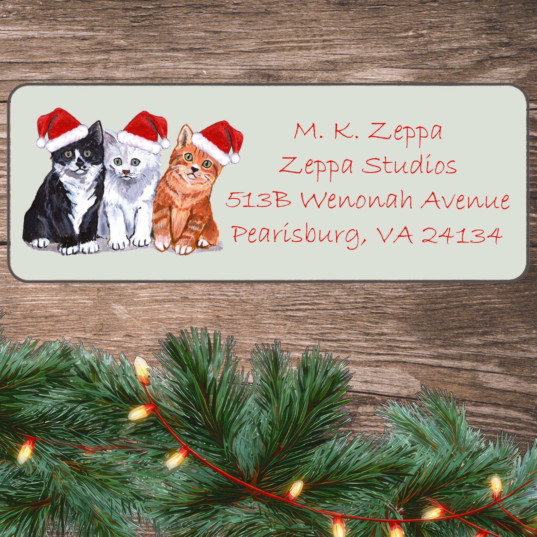 3 Kittens in Santa Hats on an ivory return address label set off by a wood surface with holiday greenery background.