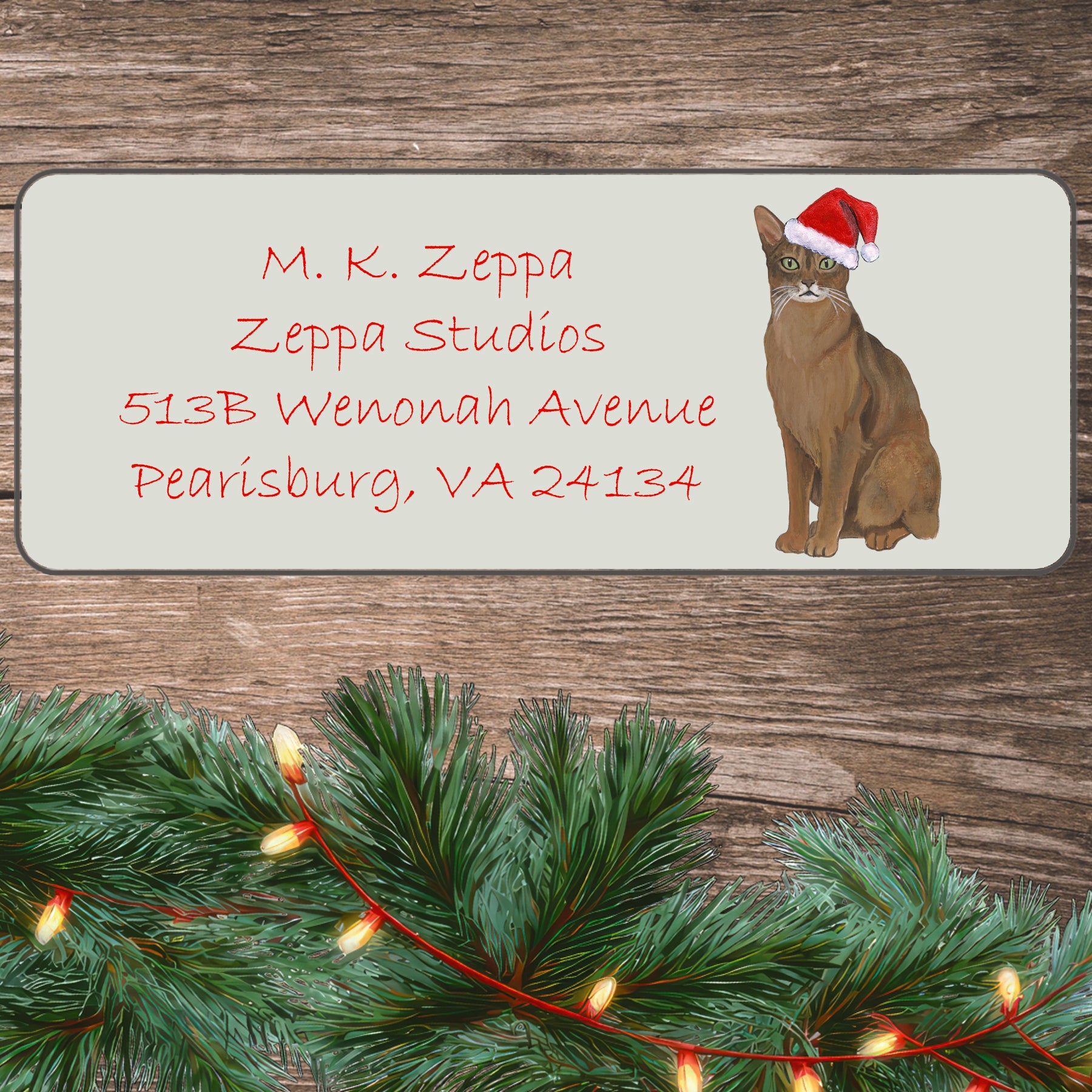 An illustration of an Abyssinian Cat wearing a Santa Hat on an ivory return address label set off by a wood surface with holiday greenery background.