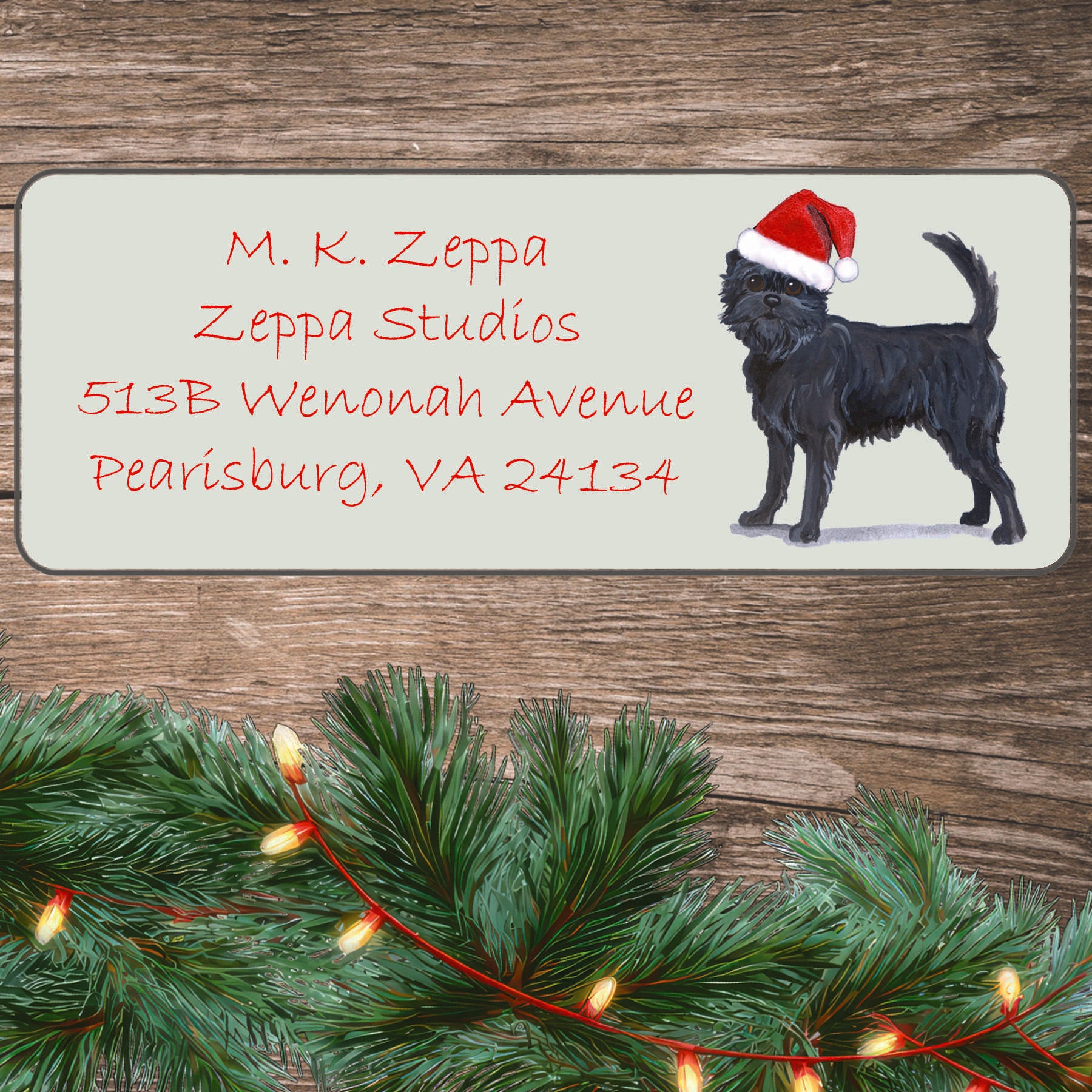 An illustration of an Affenpinscher wearing a Santa Hat on an ivory return address label set off by a wood surface with holiday greenery background.