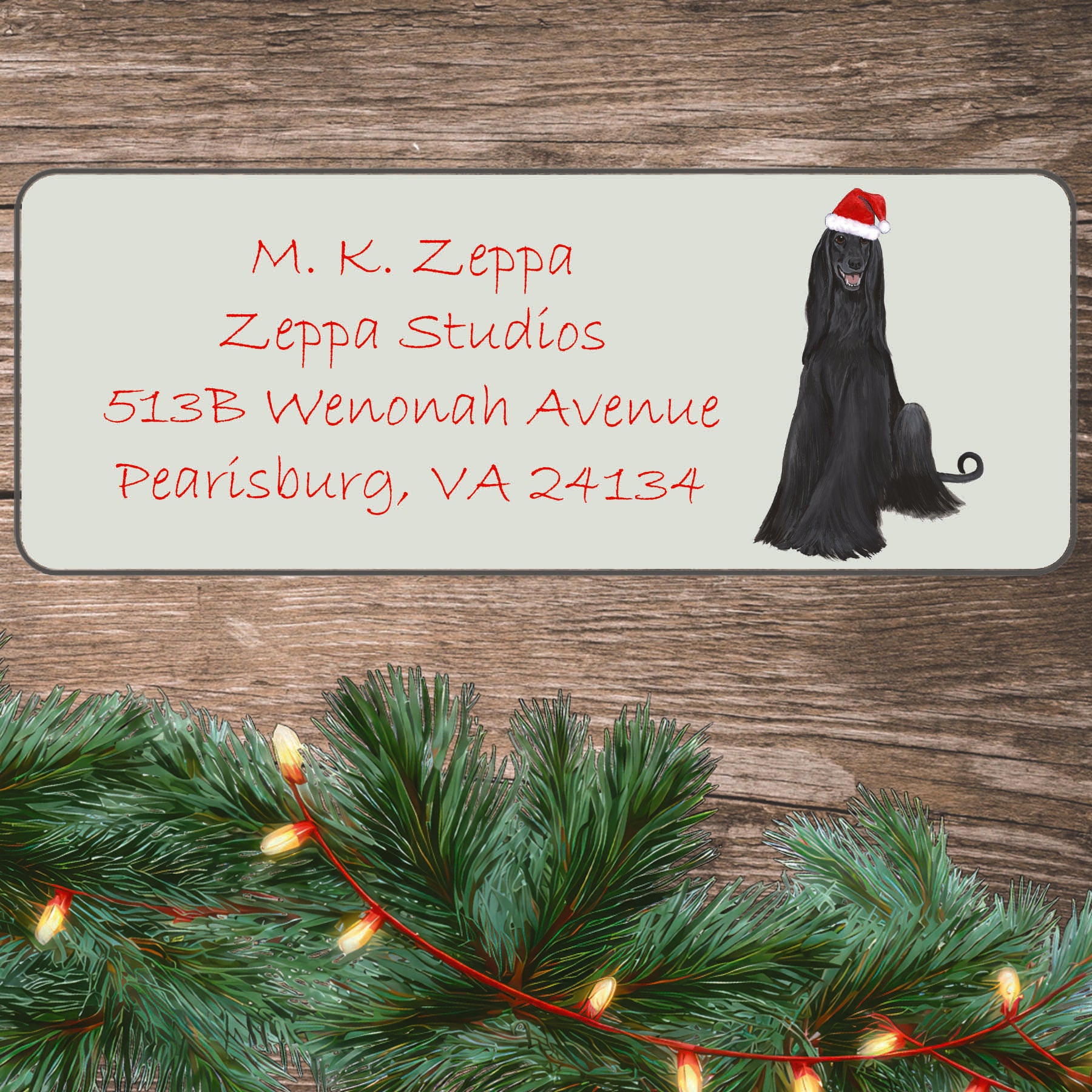 An illustration of a black Afghan Hound wearing a Santa Hat on an ivory return address label set off by a wood surface with holiday greenery background.