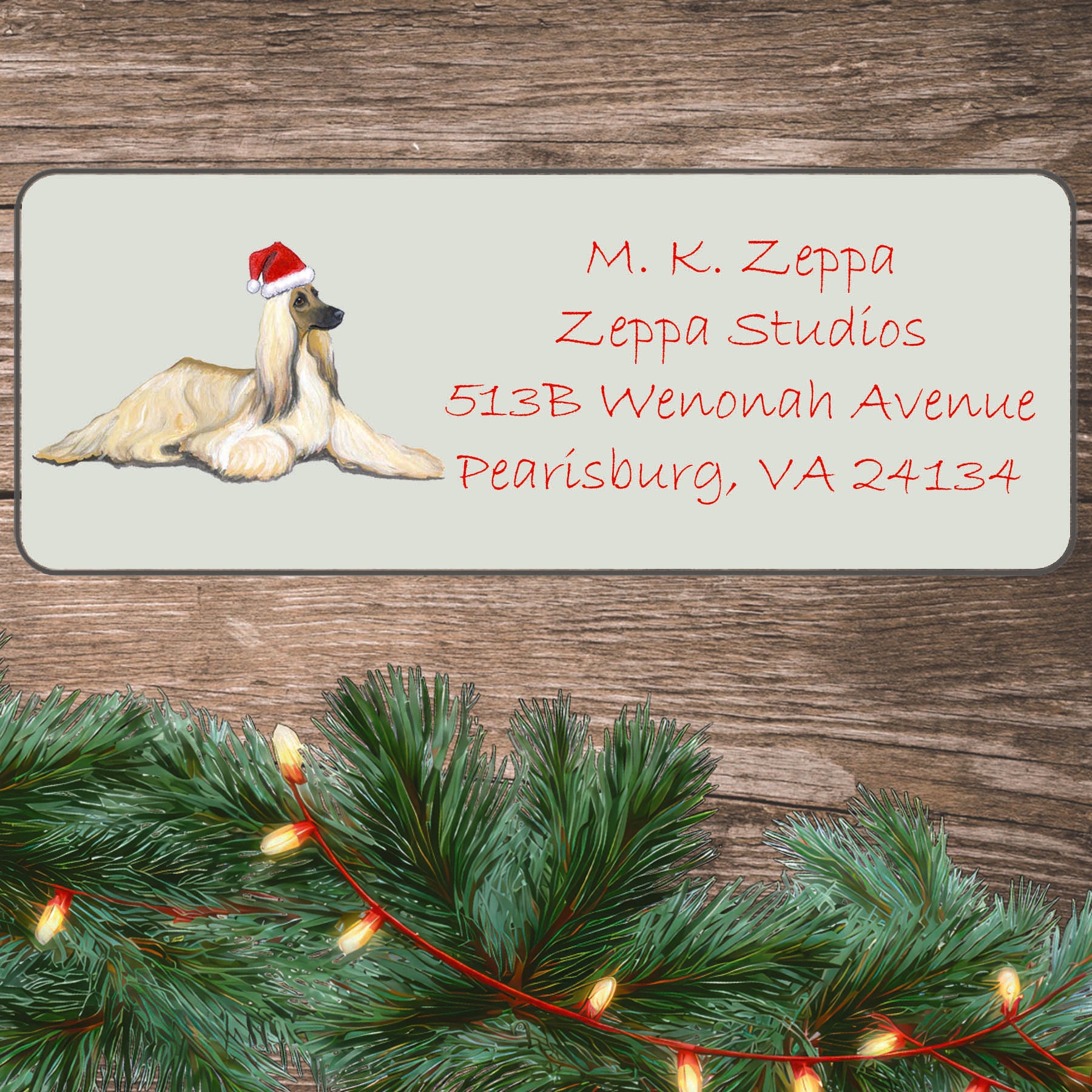 An illustration of an Afghan Hound wearing a Santa Hat on an ivory return address label set off by a wood surface with holiday greenery background.
