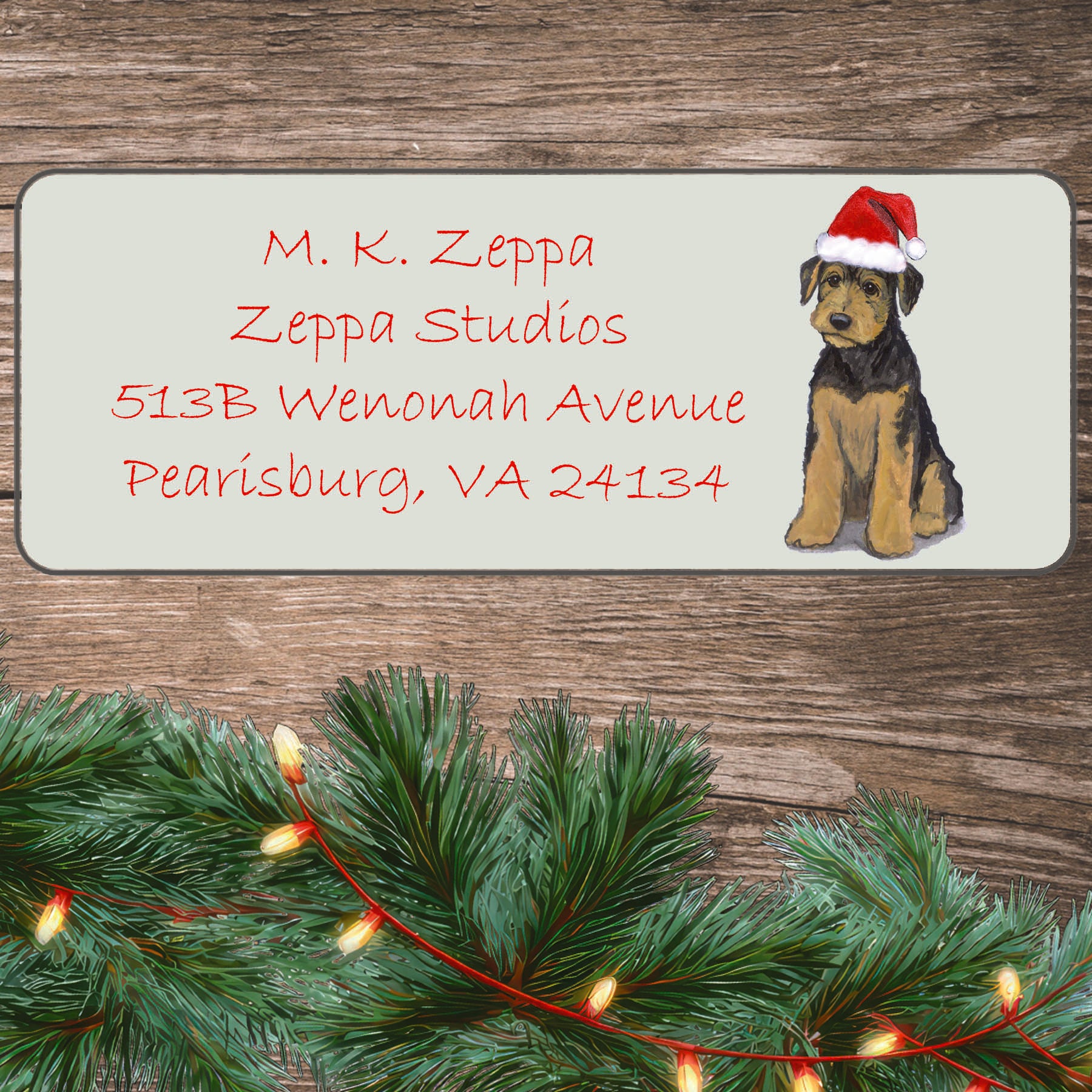 An illustration of an Airedale Terrier puppy wearing a Santa Hat on an ivory return address label set off by a wood surface with holiday greenery background.