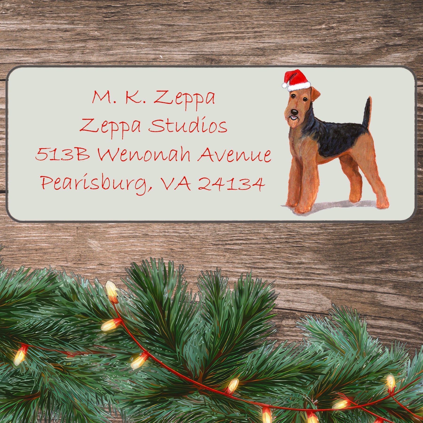 An illustration of an Airedale Terrier wearing a Santa Hat on an ivory return address label set off by a wood surface with holiday greenery background.
