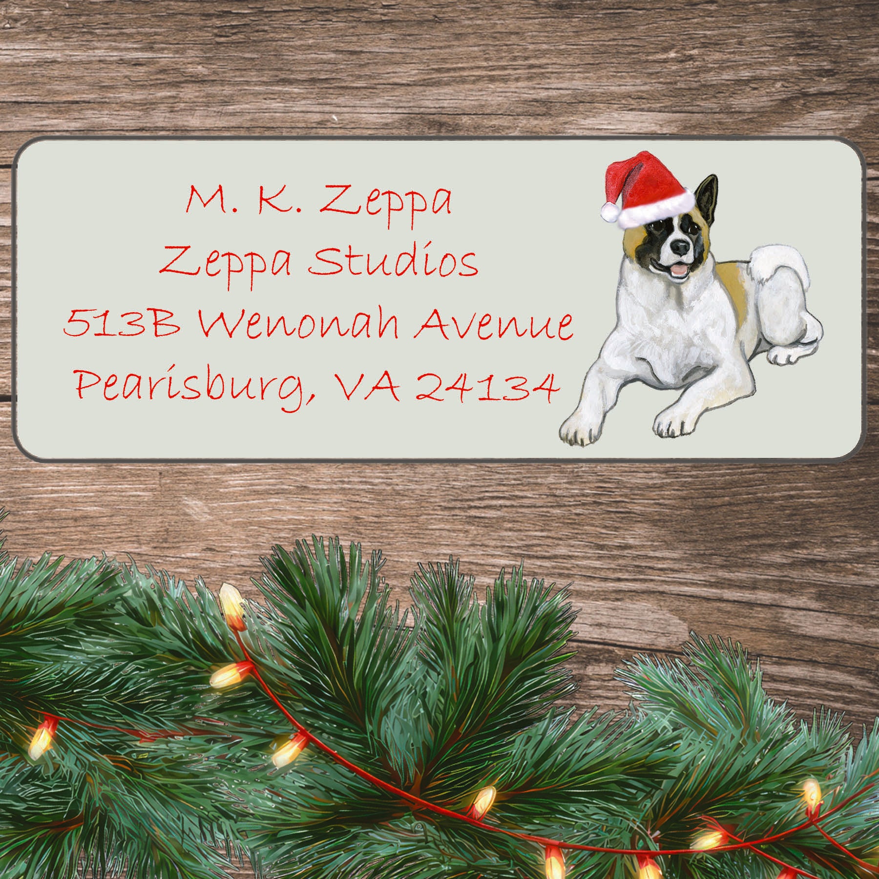 An illustration of a tan and white Akita wearing a Santa Hat on an ivory return address label set off by a wood surface with holiday greenery background.