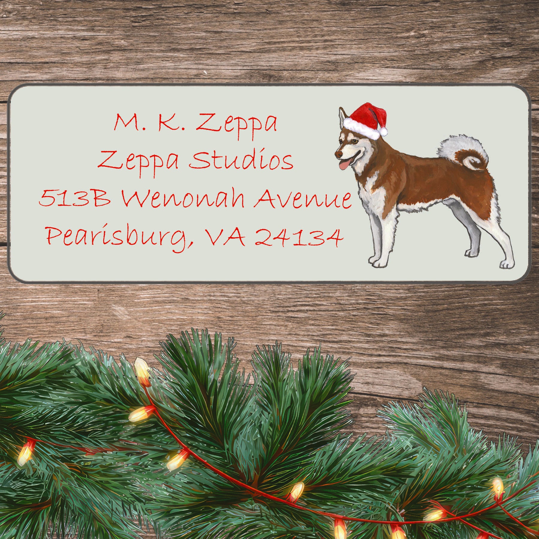 An Alaskan Klee Kai in a Santa Hat illustration on an ivory return address label set off by a wood surface with holiday greenery background.
