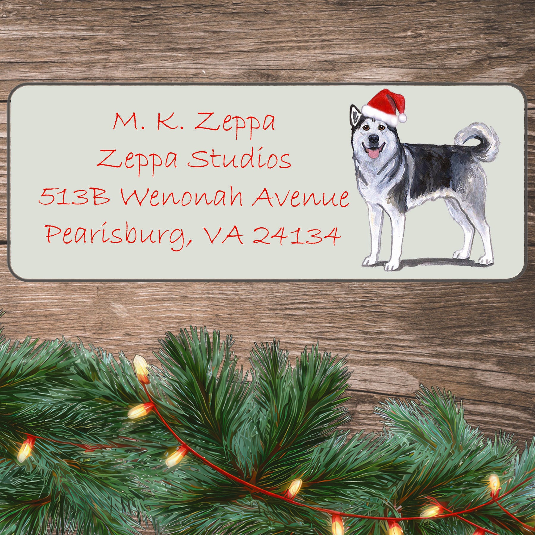 An illustration of an Alaskan Malamute wearing a Santa Hat on an ivory return address label set off by a wood surface with holiday greenery background.