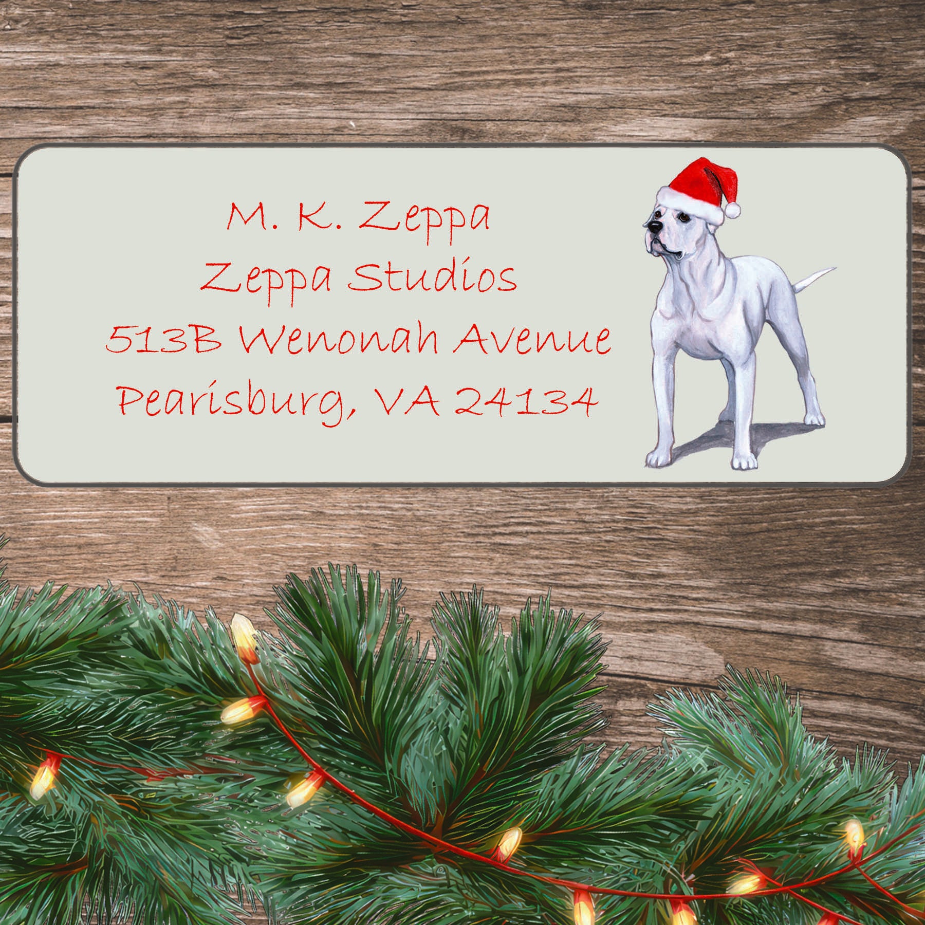 An illustration of an American Bulldog wearing a Santa Hat on an ivory return address label set off by a wood surface with holiday greenery background.