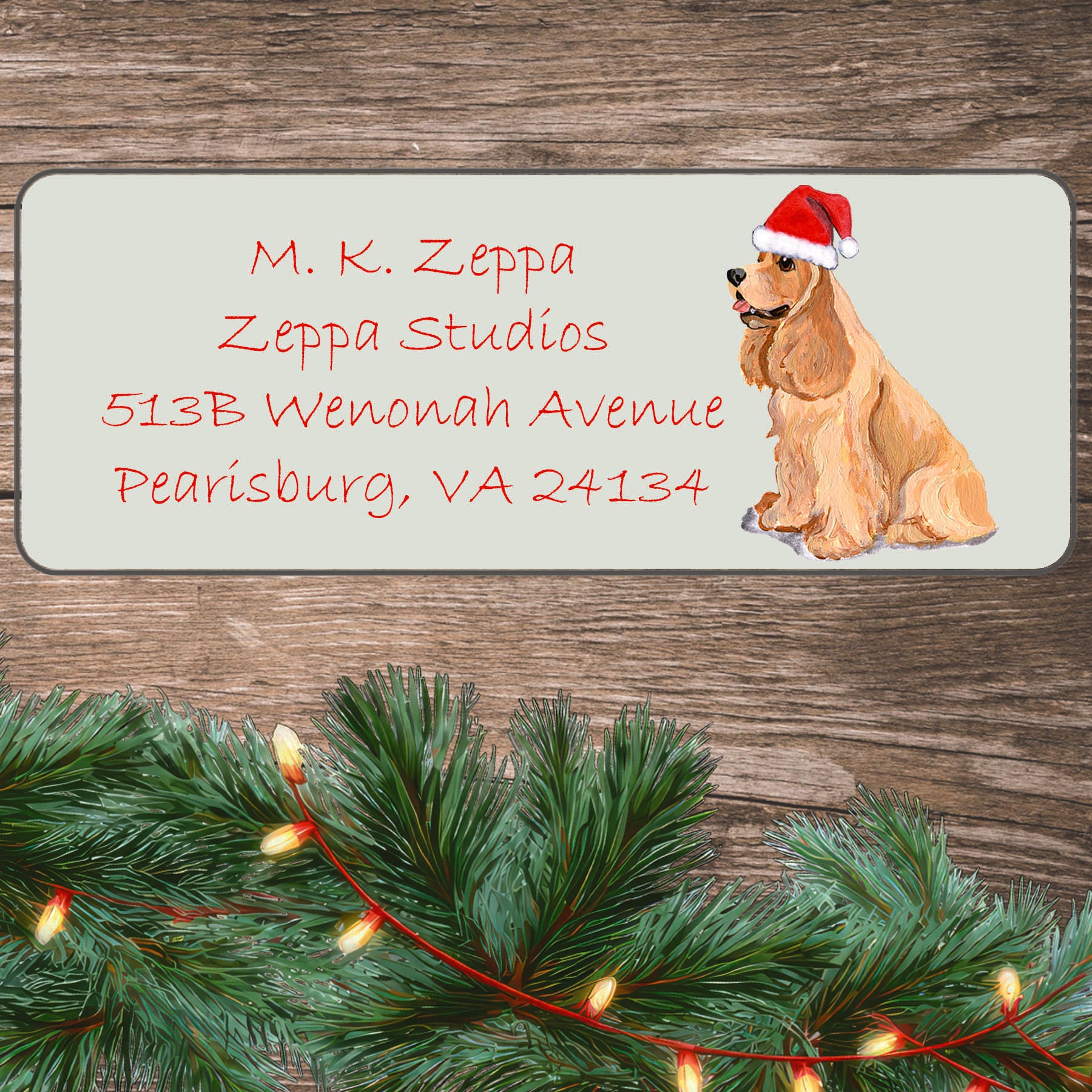 An illustration of a blonde Cocker Spaniel wearing a Santa Hat on an ivory return address label set off by a wood surface with holiday greenery background.
