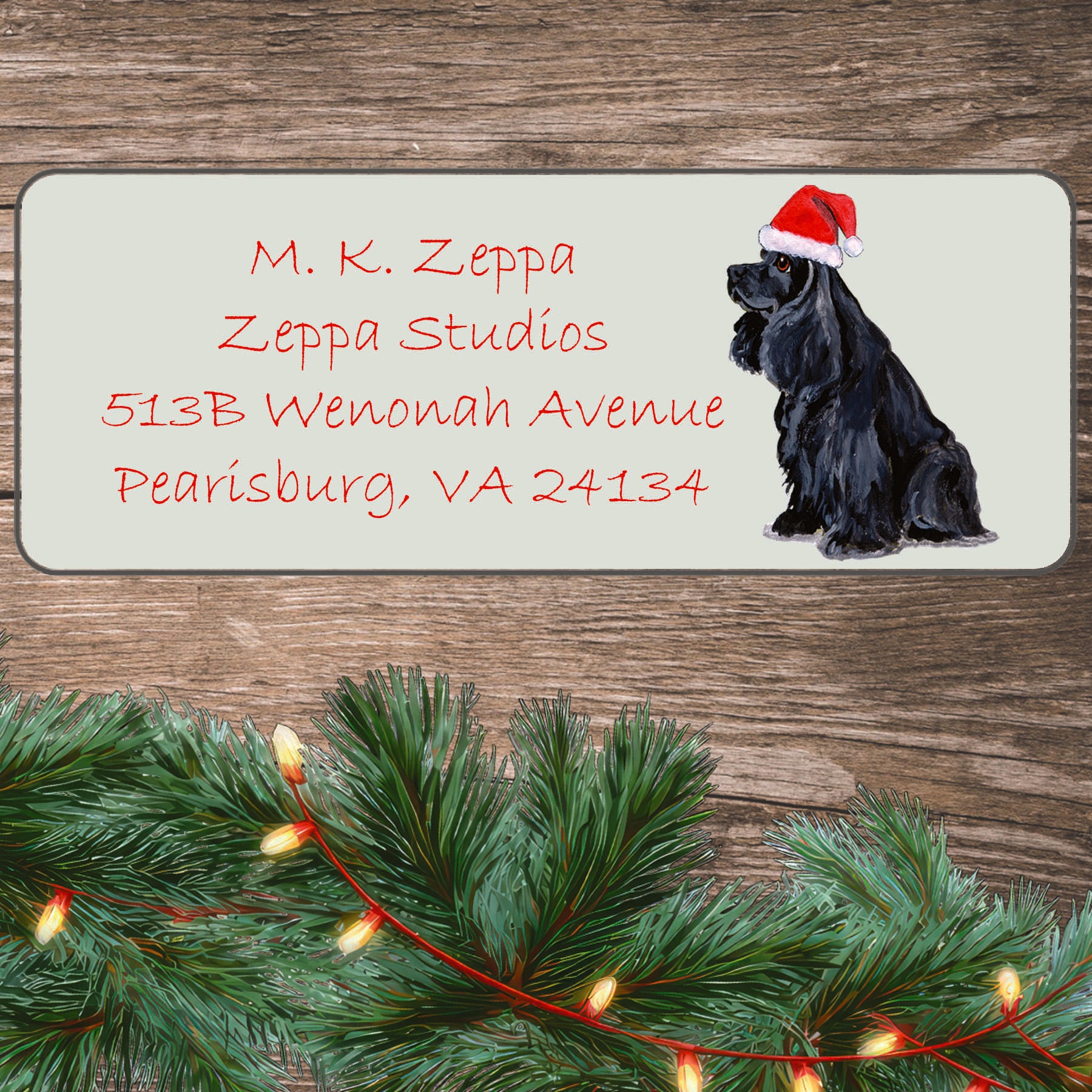 An illustration of a black Cocker Spaniel wearing a Santa Hat on an ivory return address label set off by a wood surface with holiday greenery background.