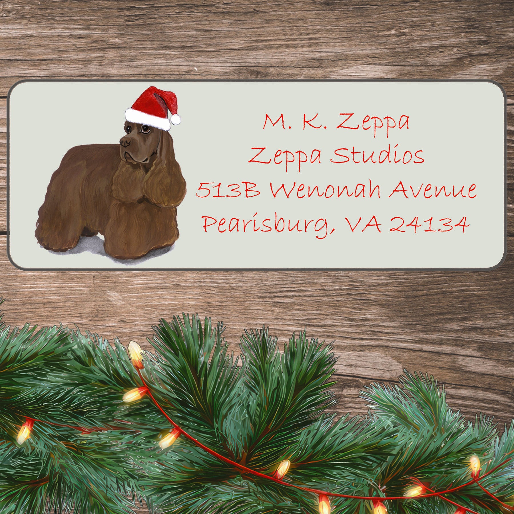 An illustration of a chocolate Cocker Spaniel wearing a Santa Hat on an ivory return address label set off by a wood surface with holiday greenery background.