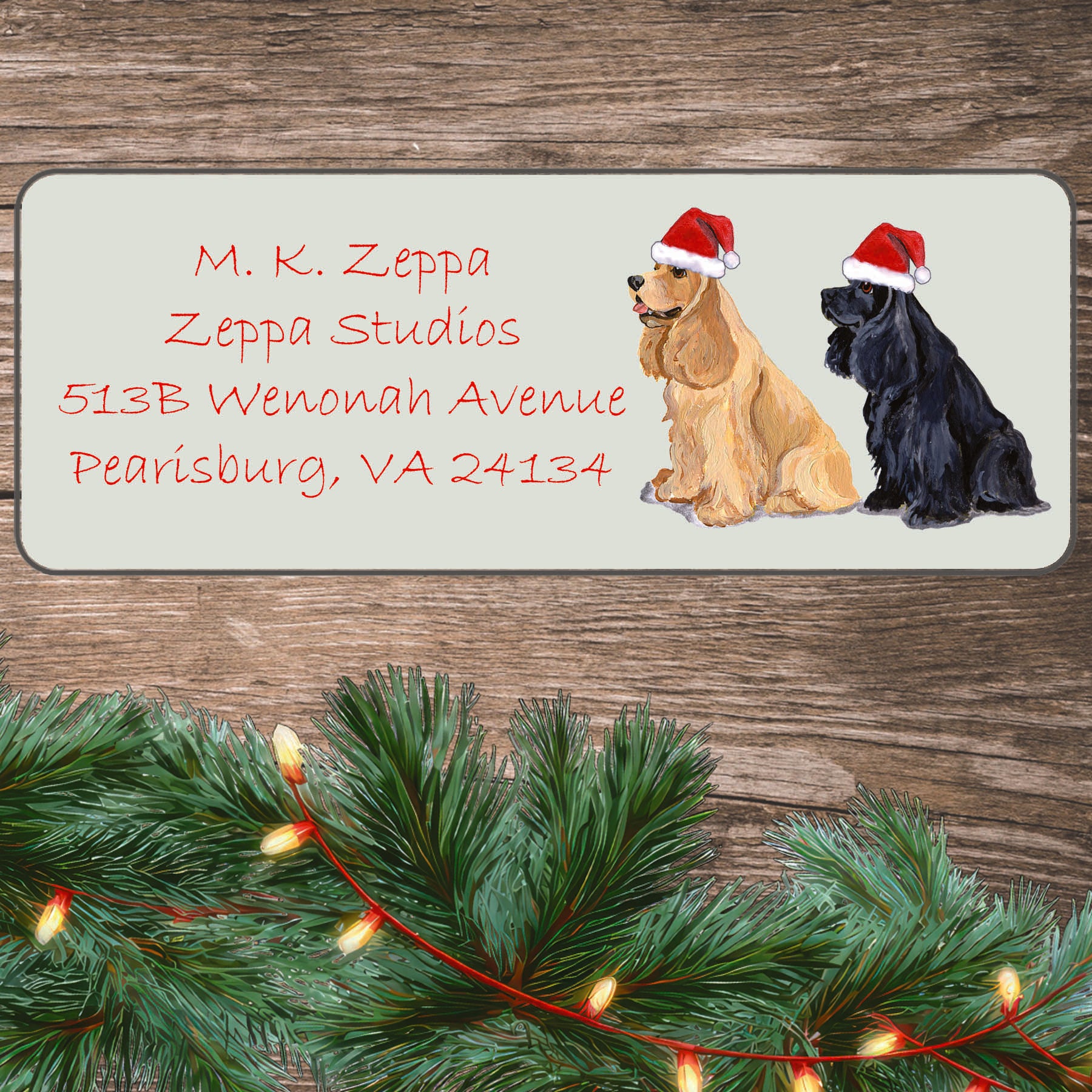 An illustration of a pair of Cocker Spaniels (tan & balck) wearing Santa Hats on an ivory return address label set off by a wood surface with holiday greenery background.