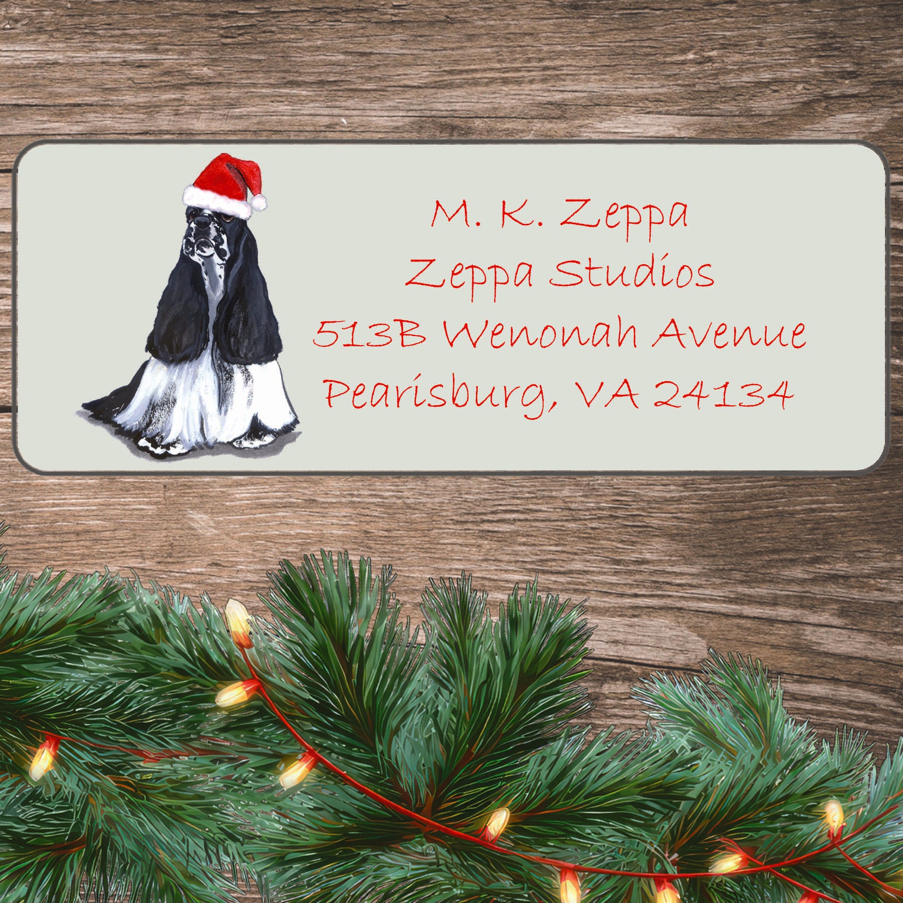 An illustration of a black and white parti Cocker Spaniel wearing a Santa Hat on an ivory return address label set off by a wood surface with holiday greenery background.