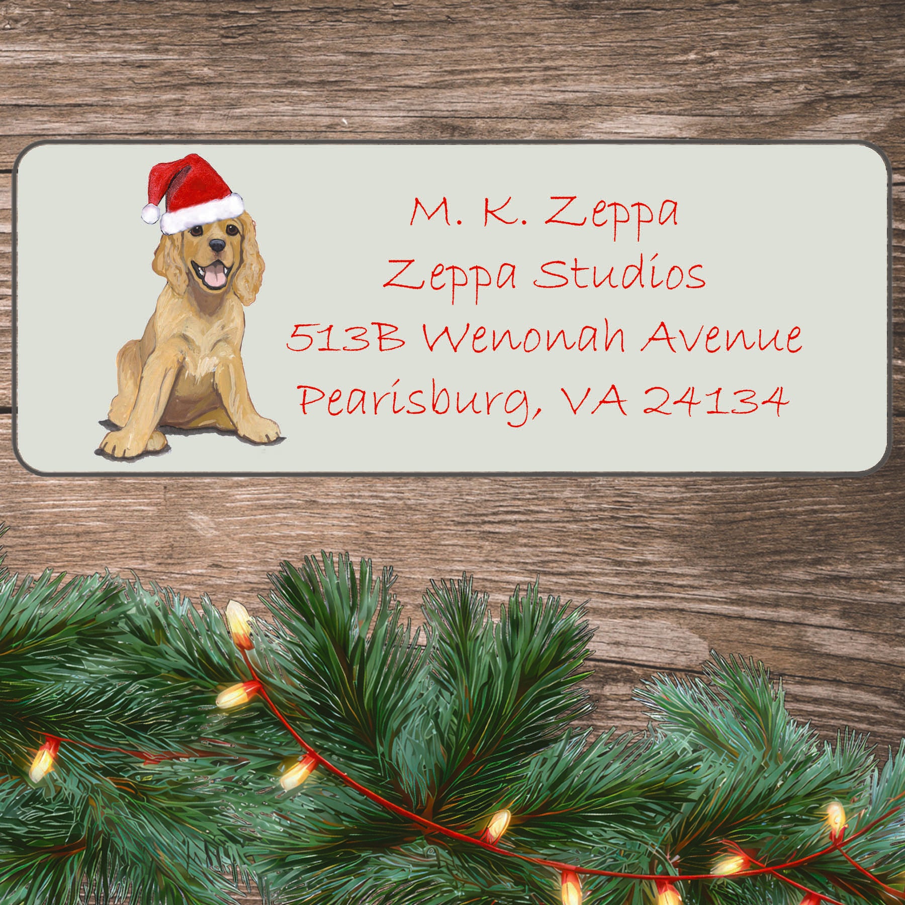 An illustration of a Cocker Spaniel puppy wearing a Santa Hat on an ivory return address label set off by a wood surface with holiday greenery background.