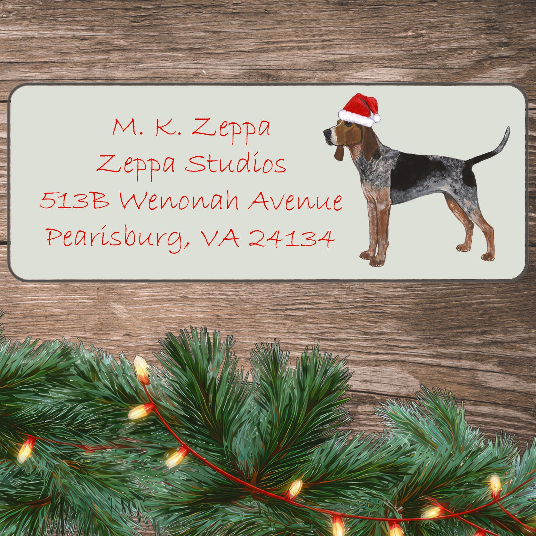 An American English Coonhound in a Santa Hat illustration on an ivory return address label set off by a wood surface with holiday greenery background.