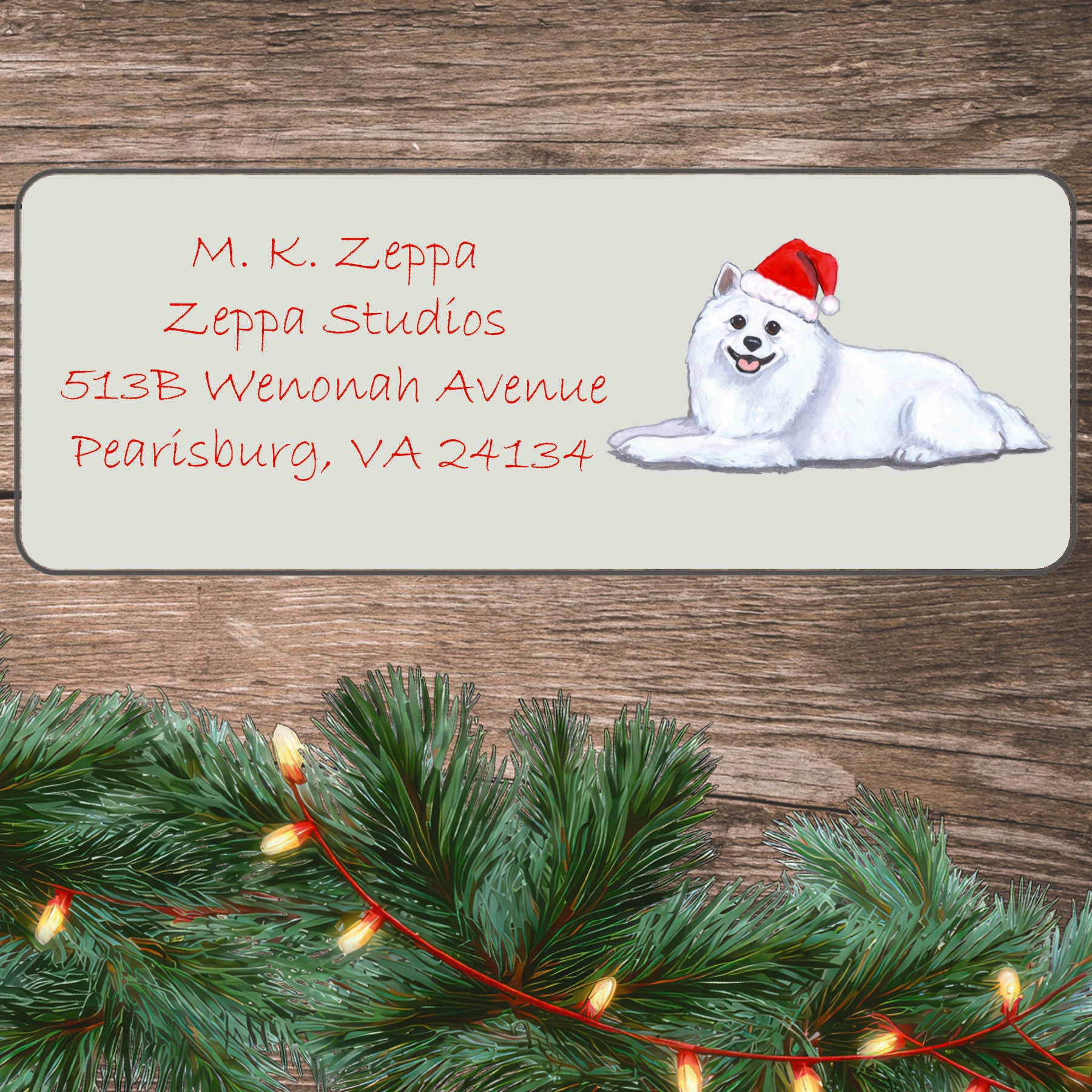 An illustration of an American Eskimo Dog wearing a Santa Hat on an ivory return address label set off by a wood surface with holiday greenery background.