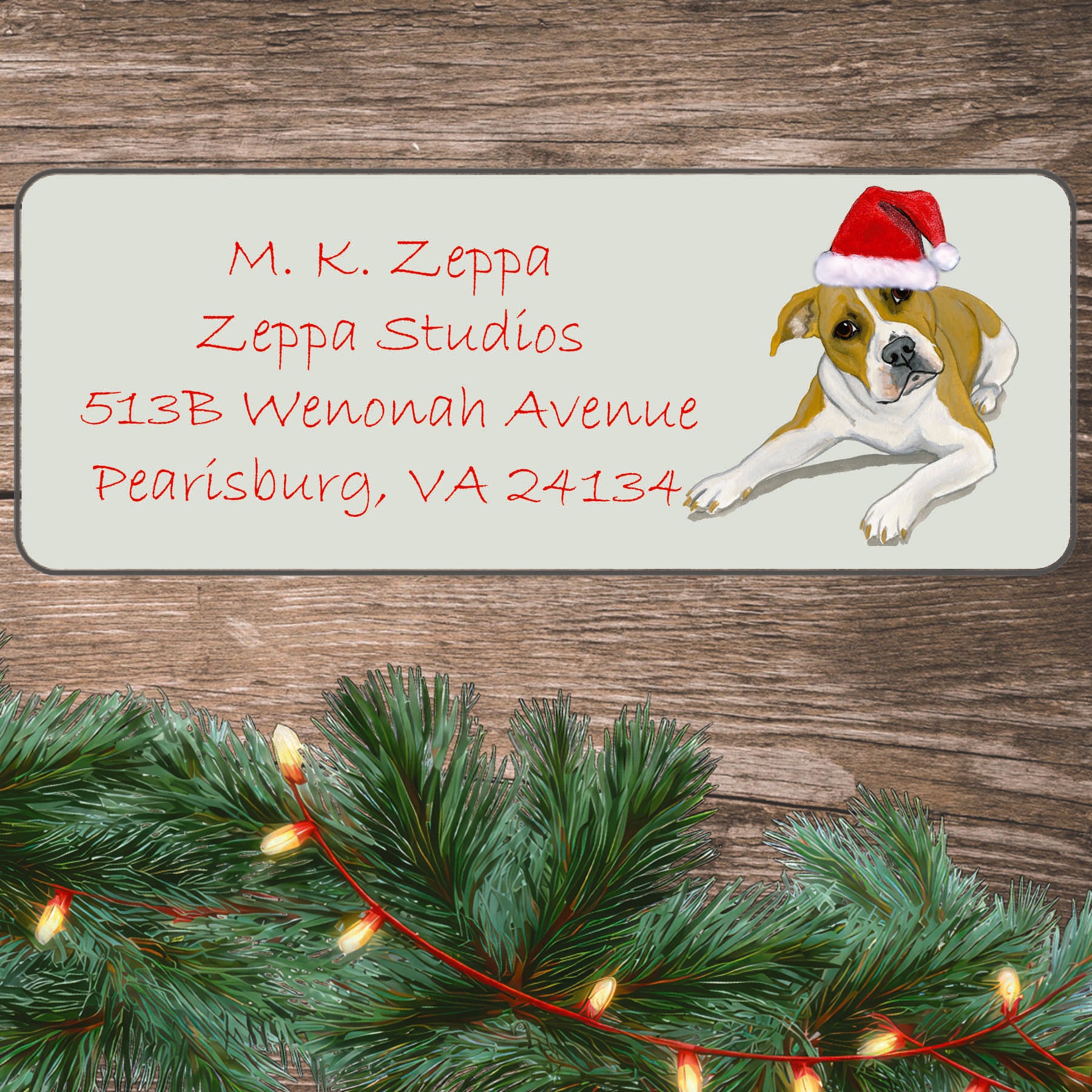 An illustration of a Pit Bull wearing a Santa Hat on an ivory return address label set off by a wood surface with holiday greenery background.