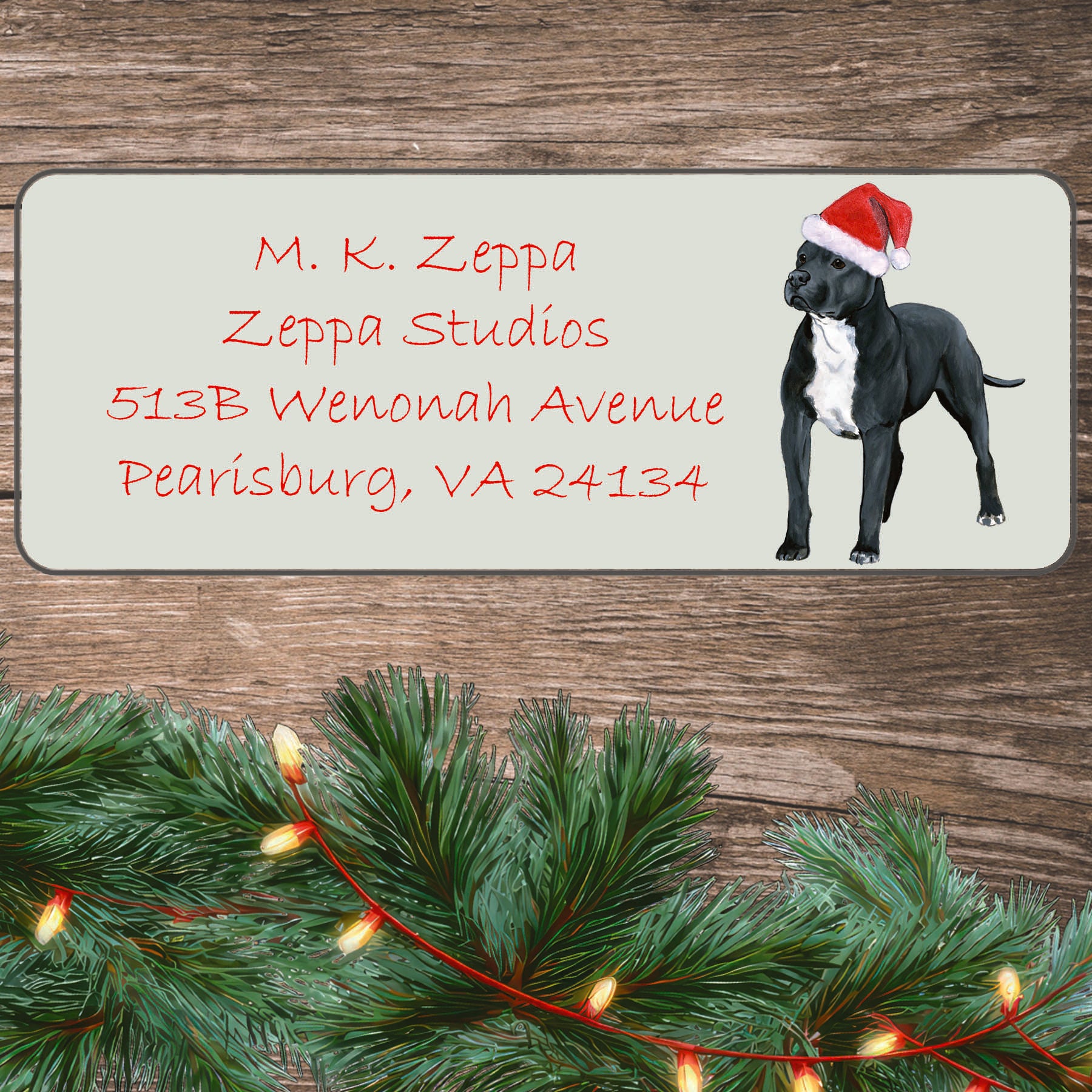 An illustration of a black and white Pit Bull wearing a Santa Hat on an ivory return address label set off by a wood surface with holiday greenery background.