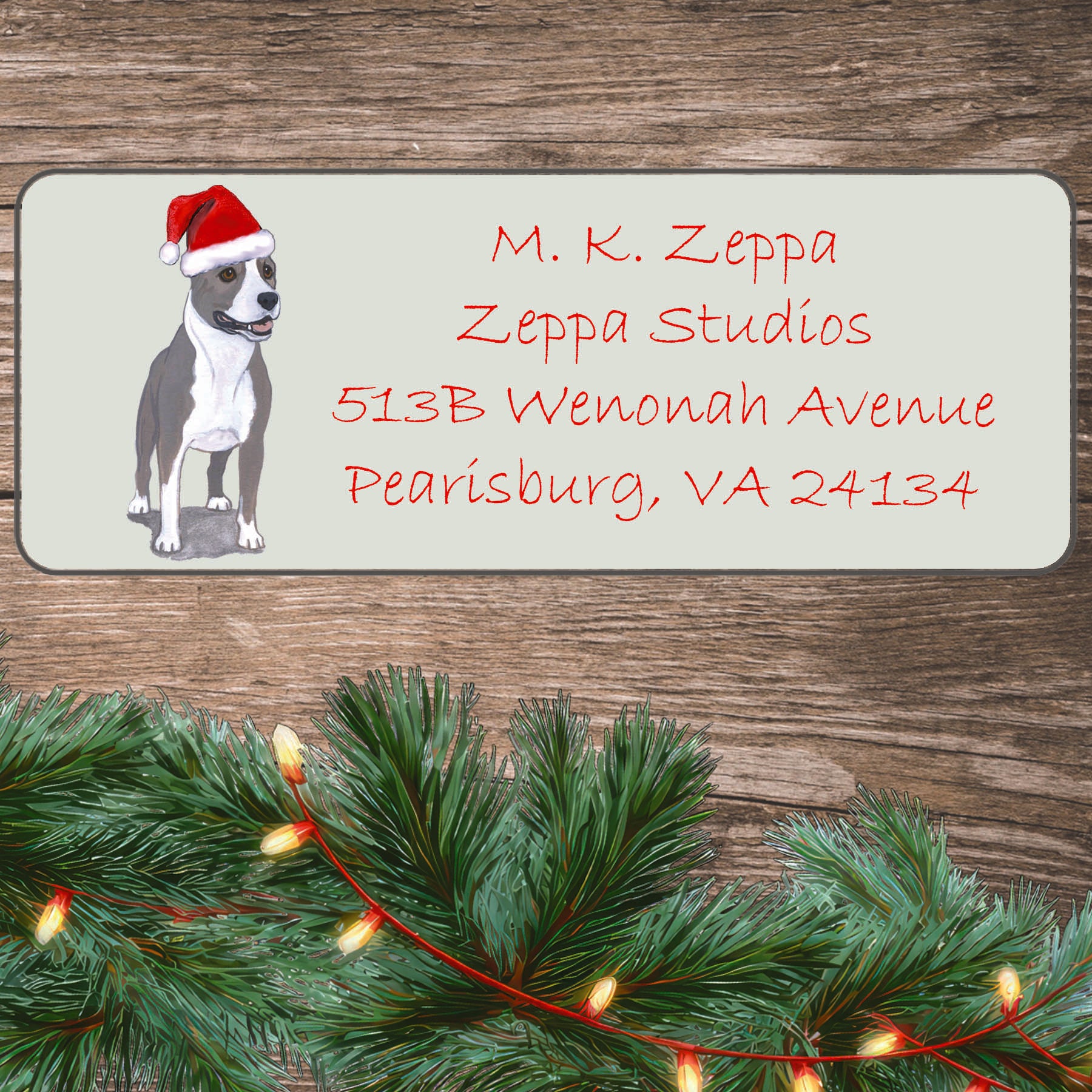An illustration of an American Staffordshire Terrier wearing a Santa Hat on an ivory return address label set off by a wood surface with holiday greenery background.