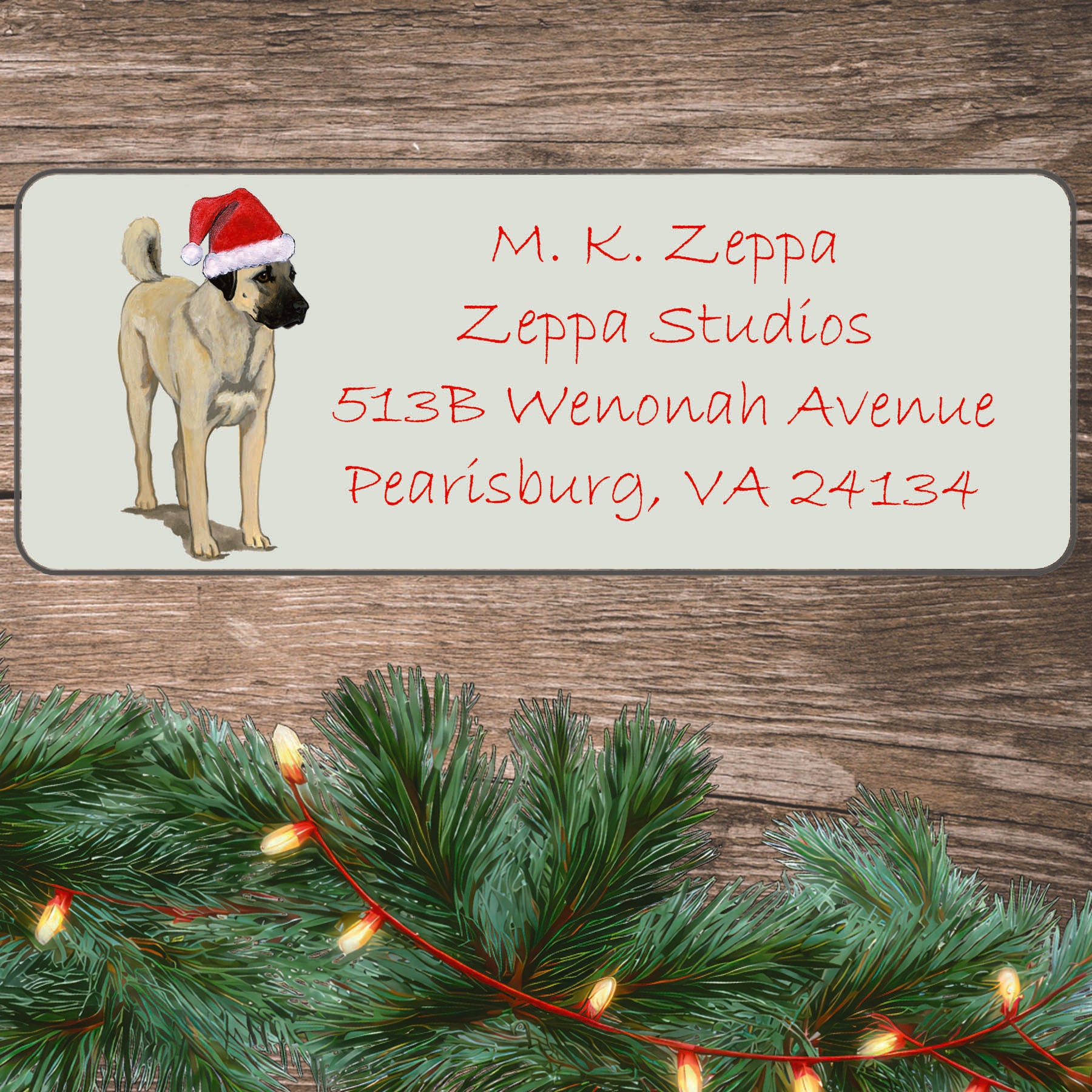 An illustration of an Anatolian Shepherd Dog wearing a Santa Hat on an ivory return address label set off by a wood surface with holiday greenery background.