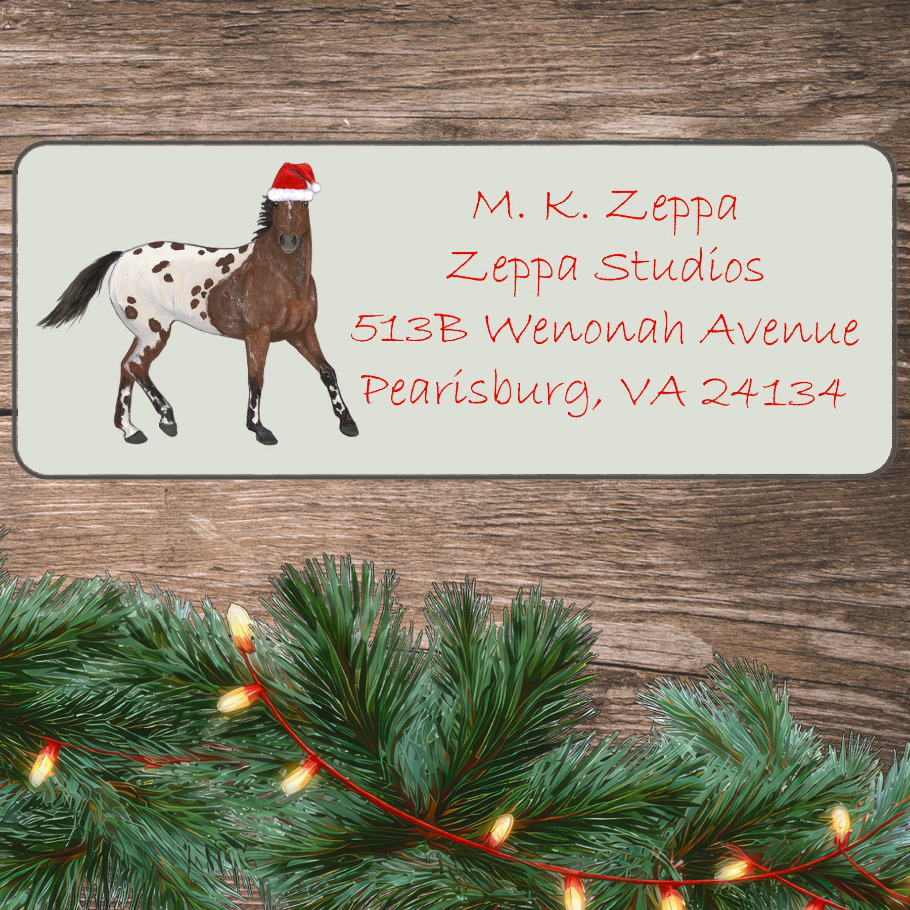 An illustration of an Appaloosa horse wearing a Santa Hat on an ivory return address label set off by a wood surface with holiday greenery background.
