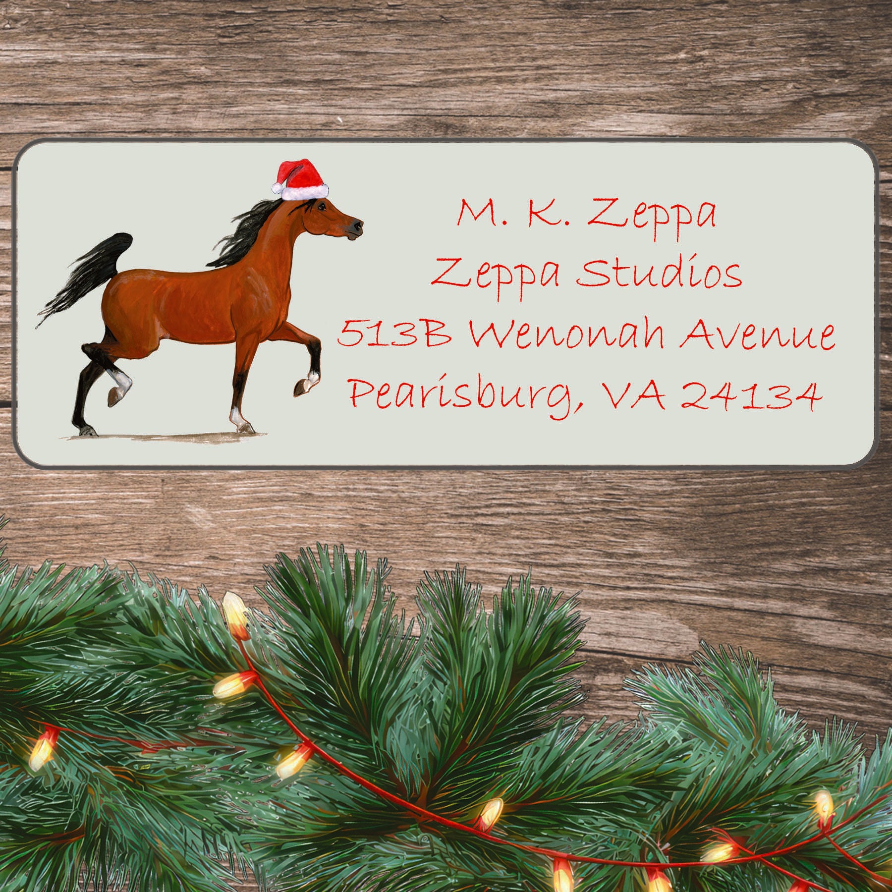 An illustration of an Arabian horse wearing a Santa Hat on an ivory return address label set off by a wood surface with holiday greenery background.