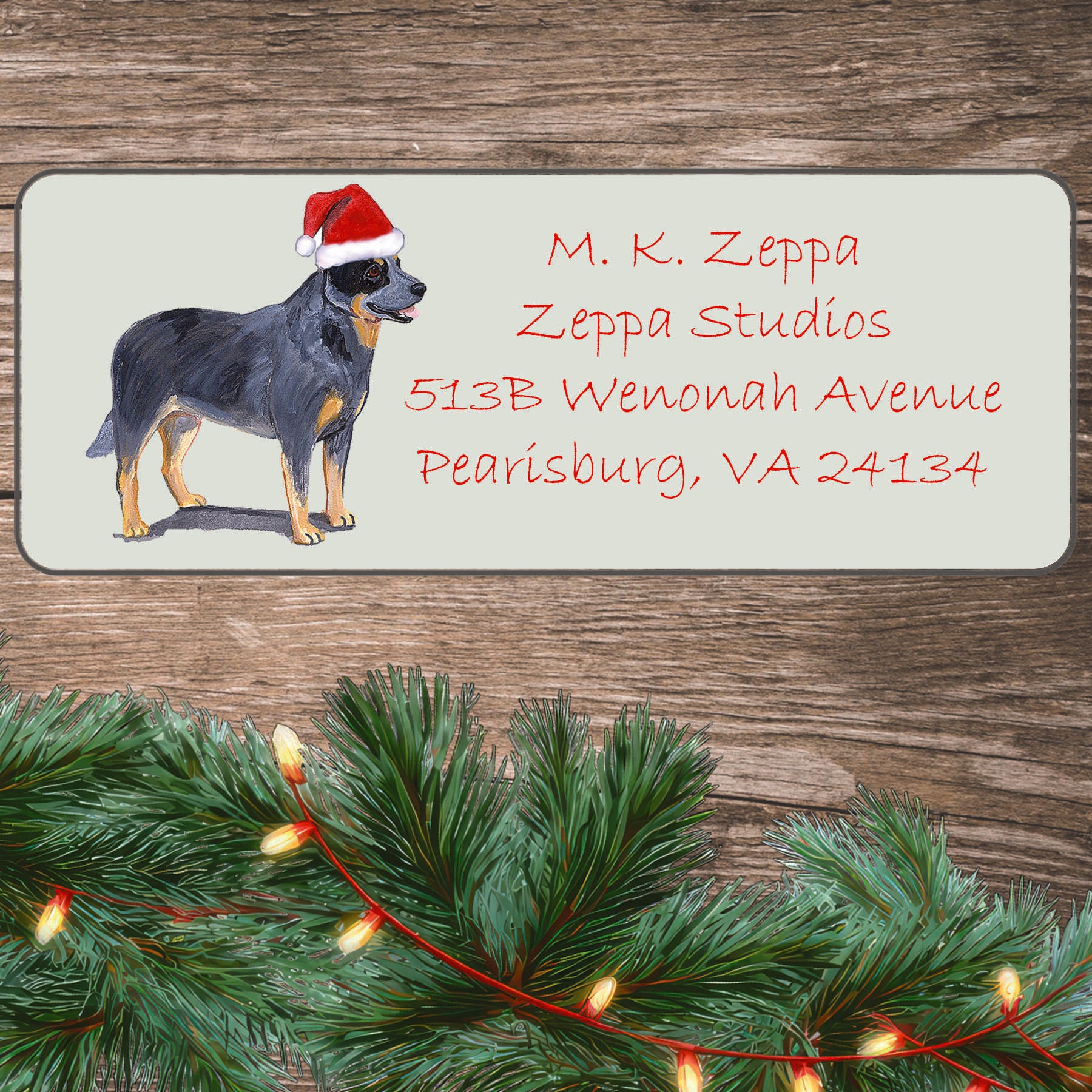 An illustration of an Australian Cattle Dog wearing a Santa Hat on an ivory return address label set off by a wood surface with holiday greenery background.