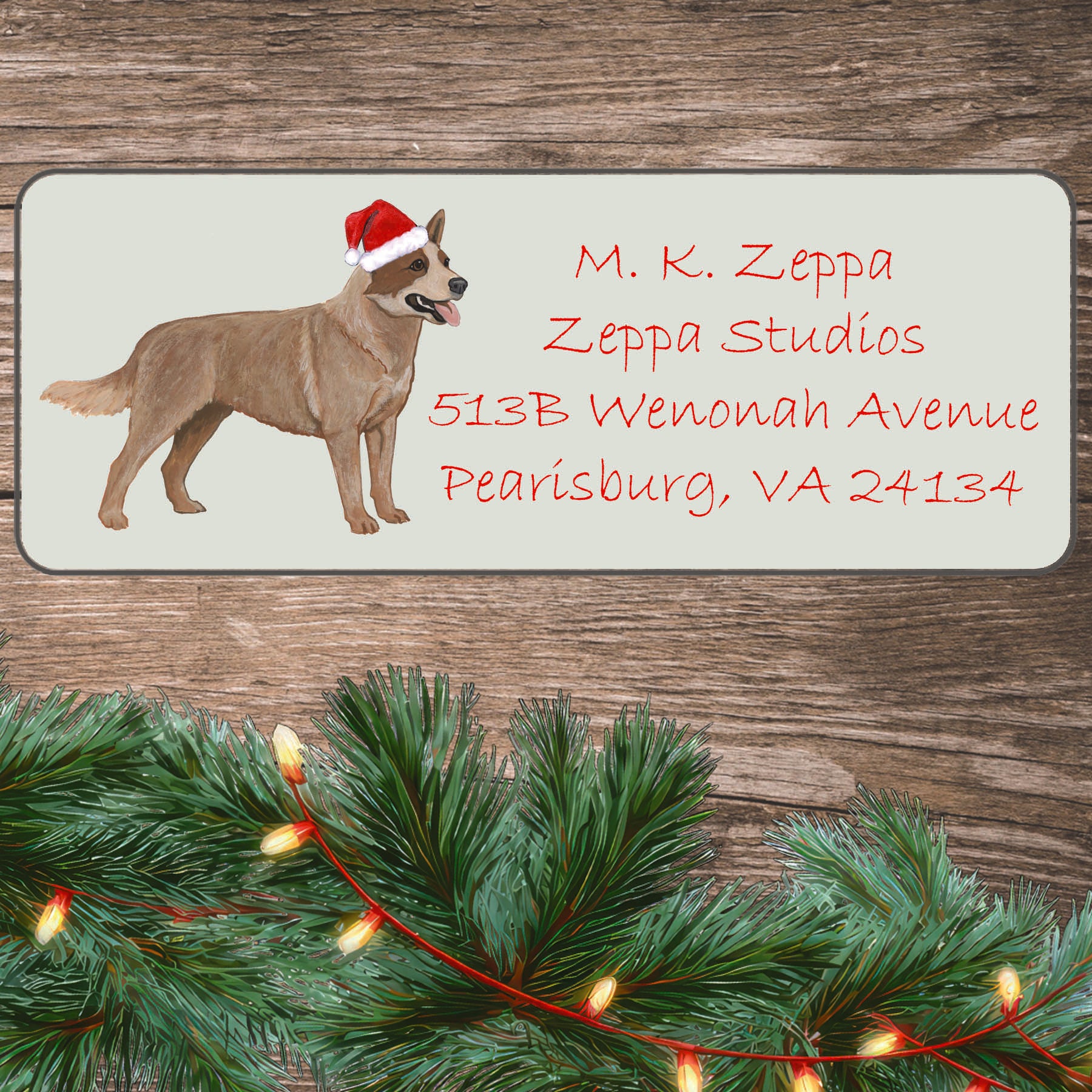An illustration of a red merle Australian Cattle Dog wearing a Santa Hat on an ivory return address label set off by a wood surface with holiday greenery background.