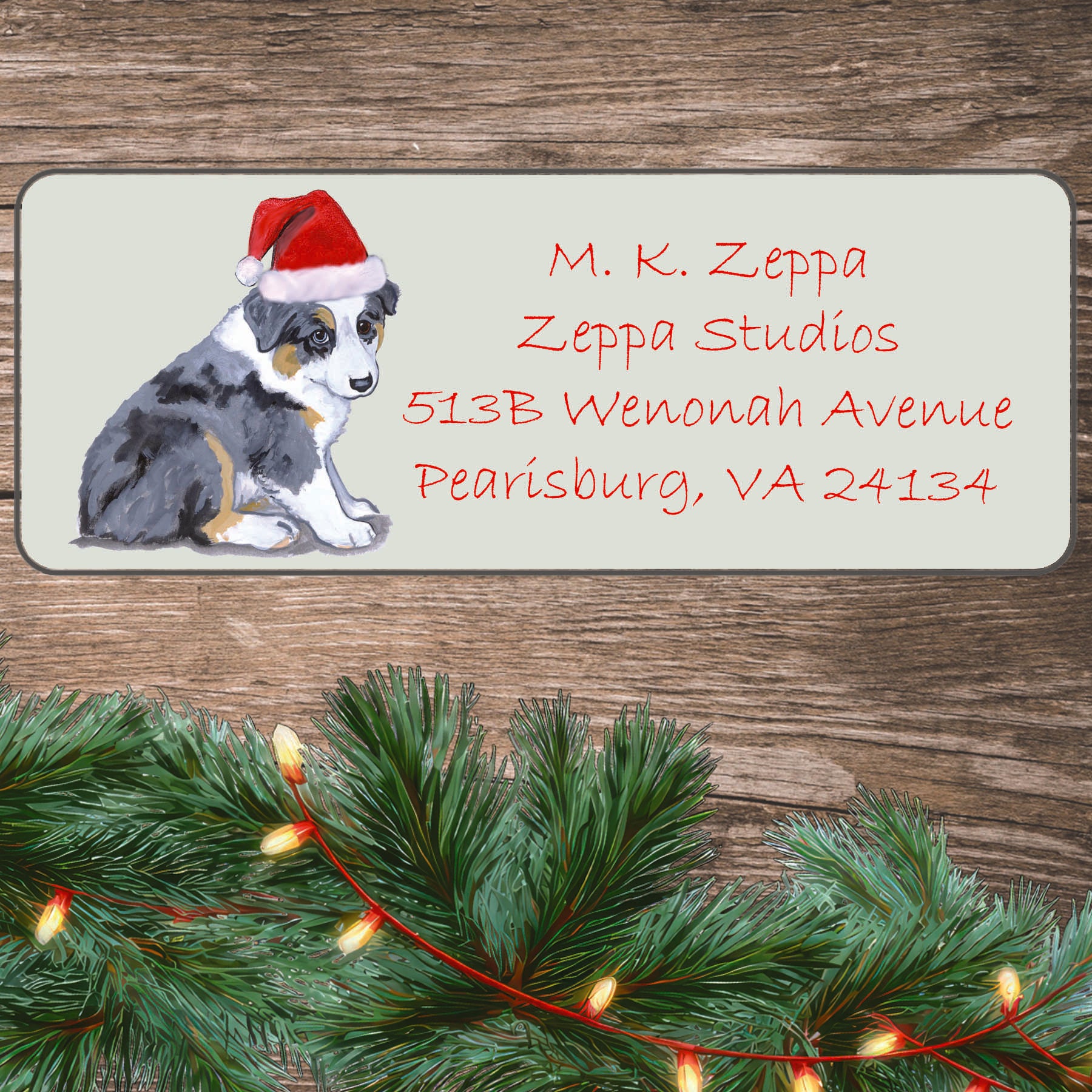An illustration of a wearing a Australian Shepherd puppy Santa Hat on an ivory return address label set off by a wood surface with holiday greenery background.