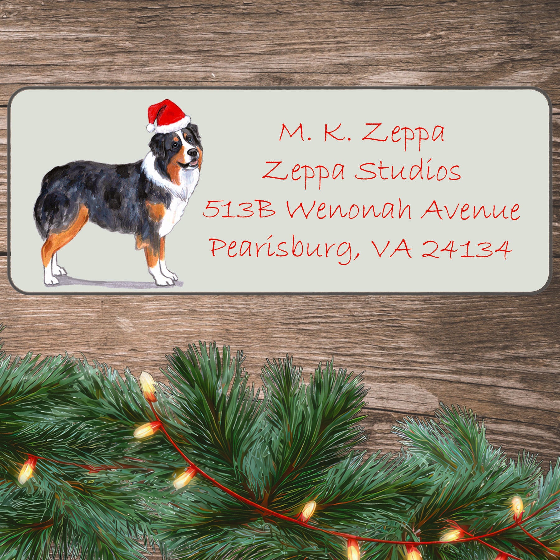An illustration of an Australian Shepherd wearing a Santa Hat on an ivory return address label set off by a wood surface with holiday greenery background.