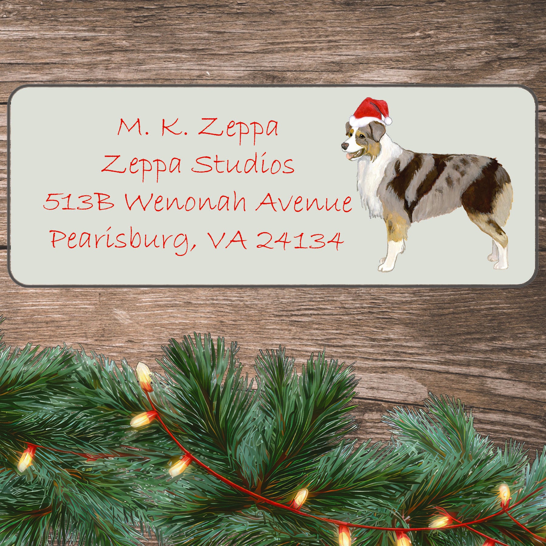 An illustration of a red merle Australian Shepherd Santa wearing a Hat on an ivory return address label set off by a wood surface with holiday greenery background.