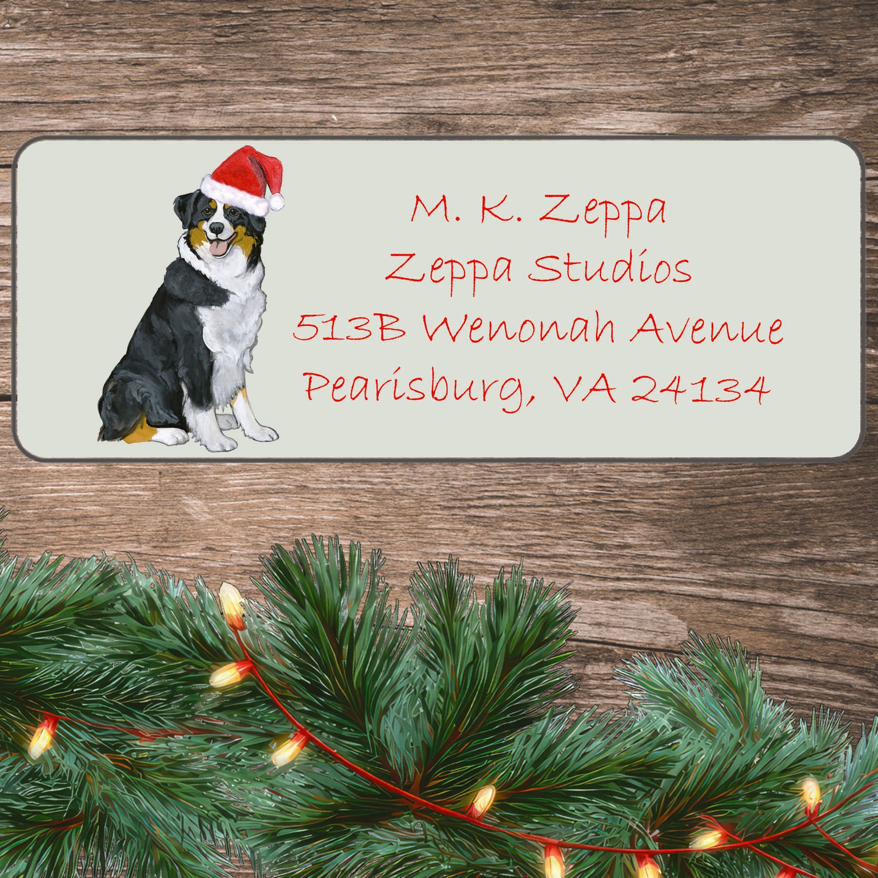 An illustration of a wearing a black tri-color Australian Shepherd Santa Hat on an ivory return address label set off by a wood surface with holiday greenery background.