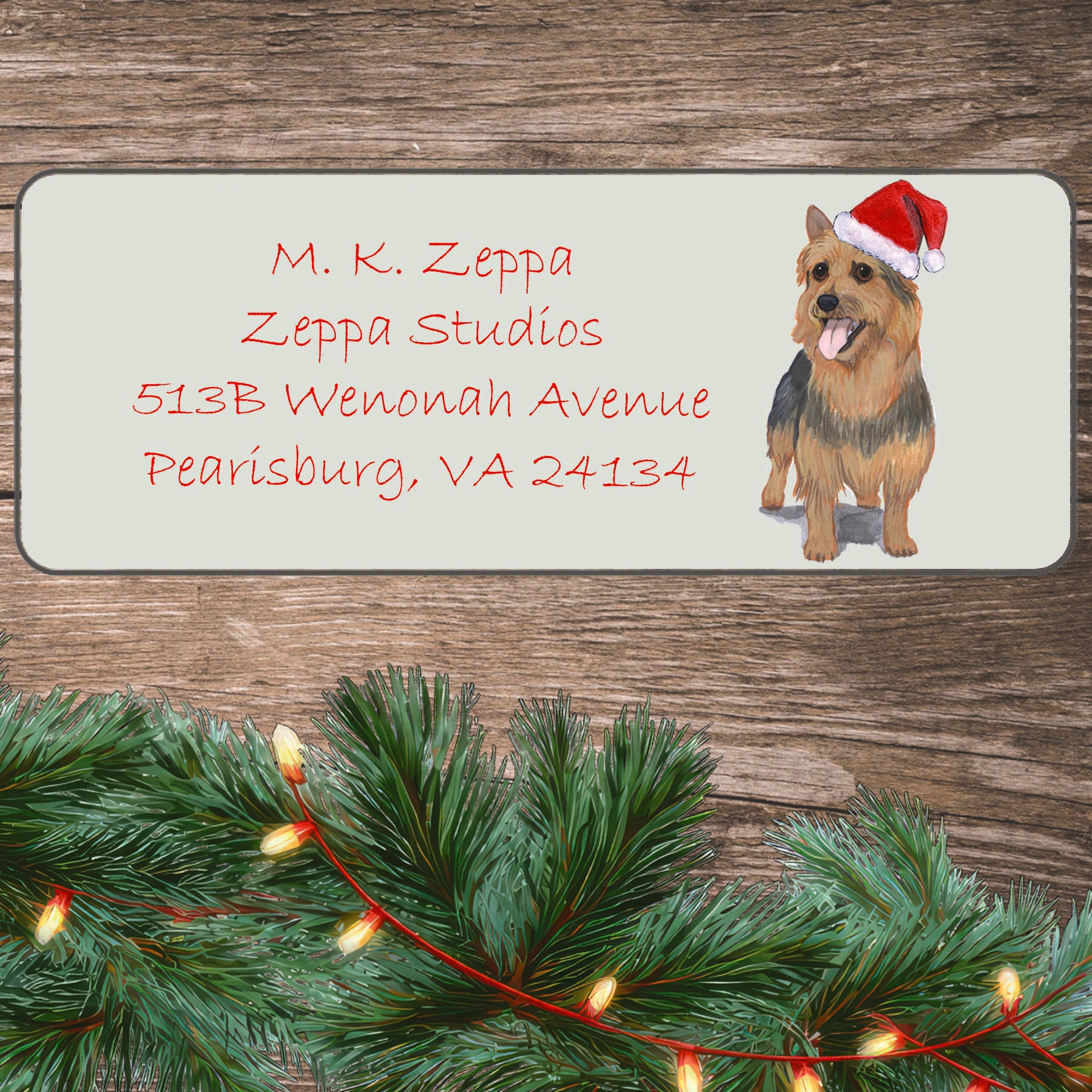 An illustration of an Australian Terrier wearing a Santa Hat on an ivory return address label set off by a wood surface with holiday greenery background.