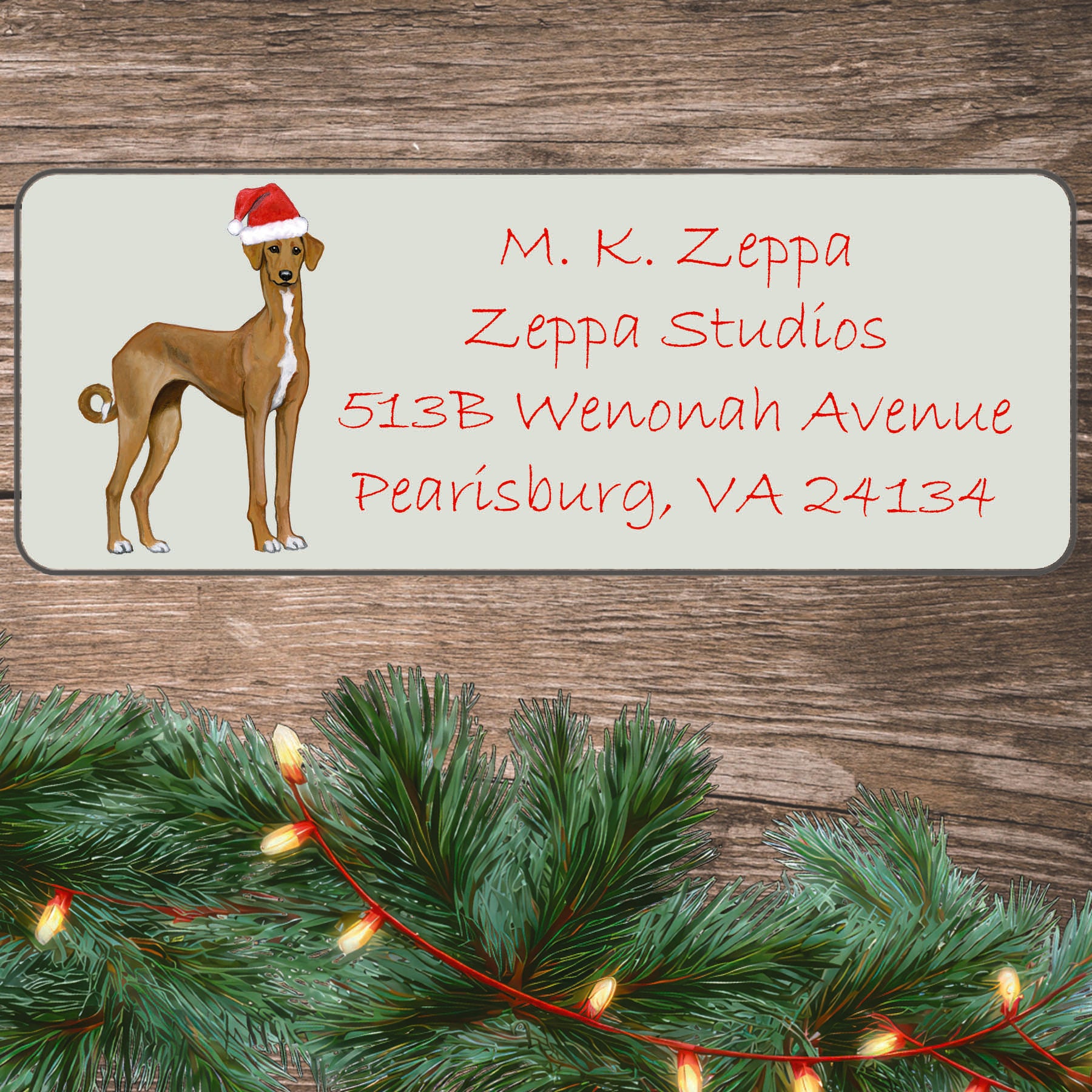 An illustration of an Azawakh wearing a Santa Hat on an ivory return address label set off by a wood surface with holiday greenery background.