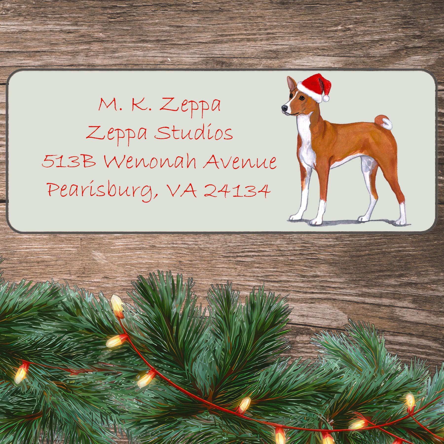 An illustration of a Basenji wearing a Santa Hat on an ivory return address label set off by a wood surface with holiday greenery background.