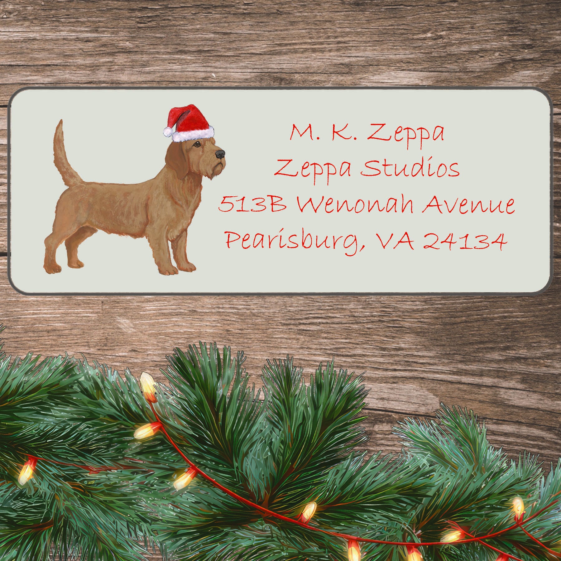 An illustration of a Basset Fauve de Bretagne wearing a Santa Hat on an ivory return address label set off by a wood surface with holiday greenery background.