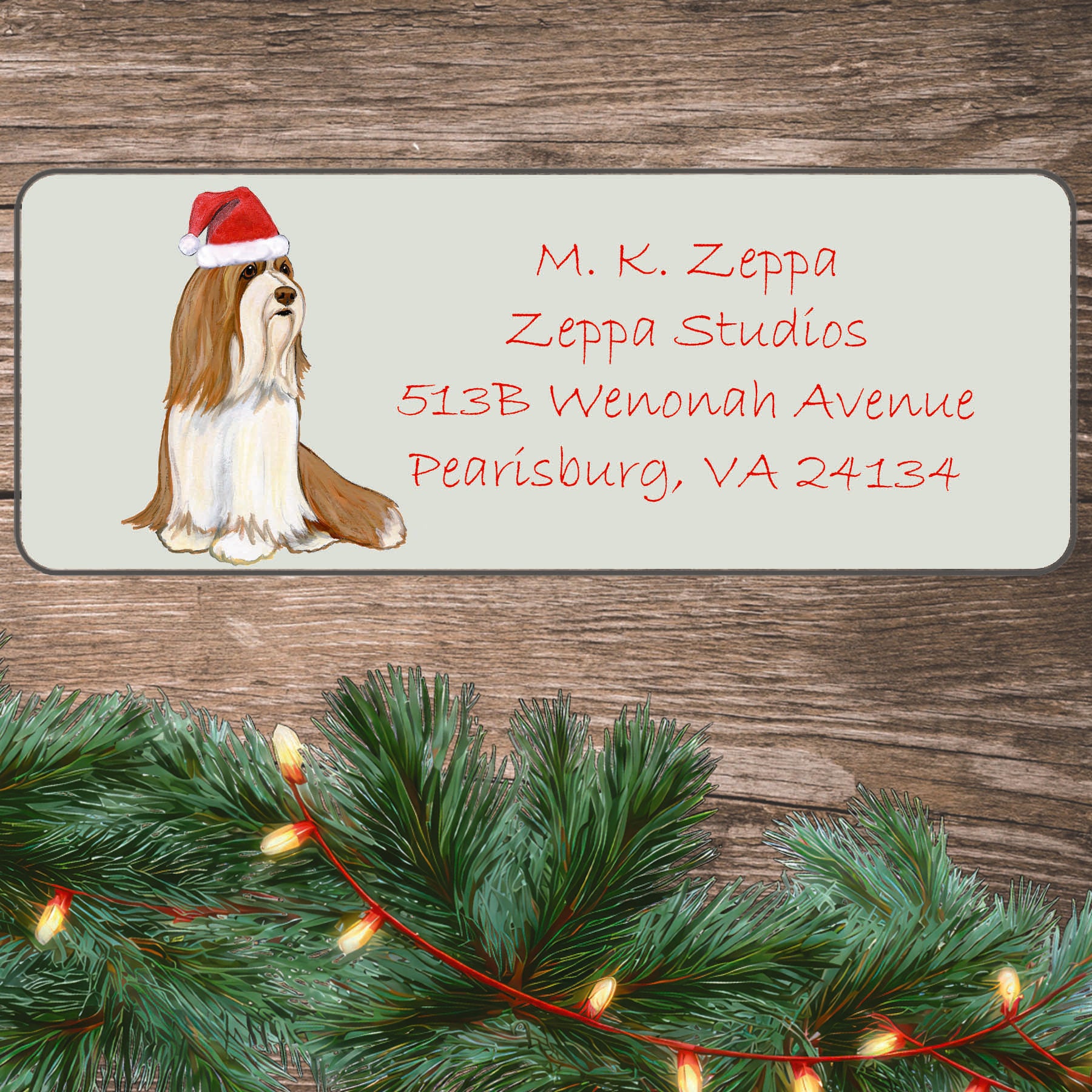 An illustration of a brown and white Bearded Collie wearing a Santa Hat on an ivory return address label set off by a wood surface with holiday greenery background.