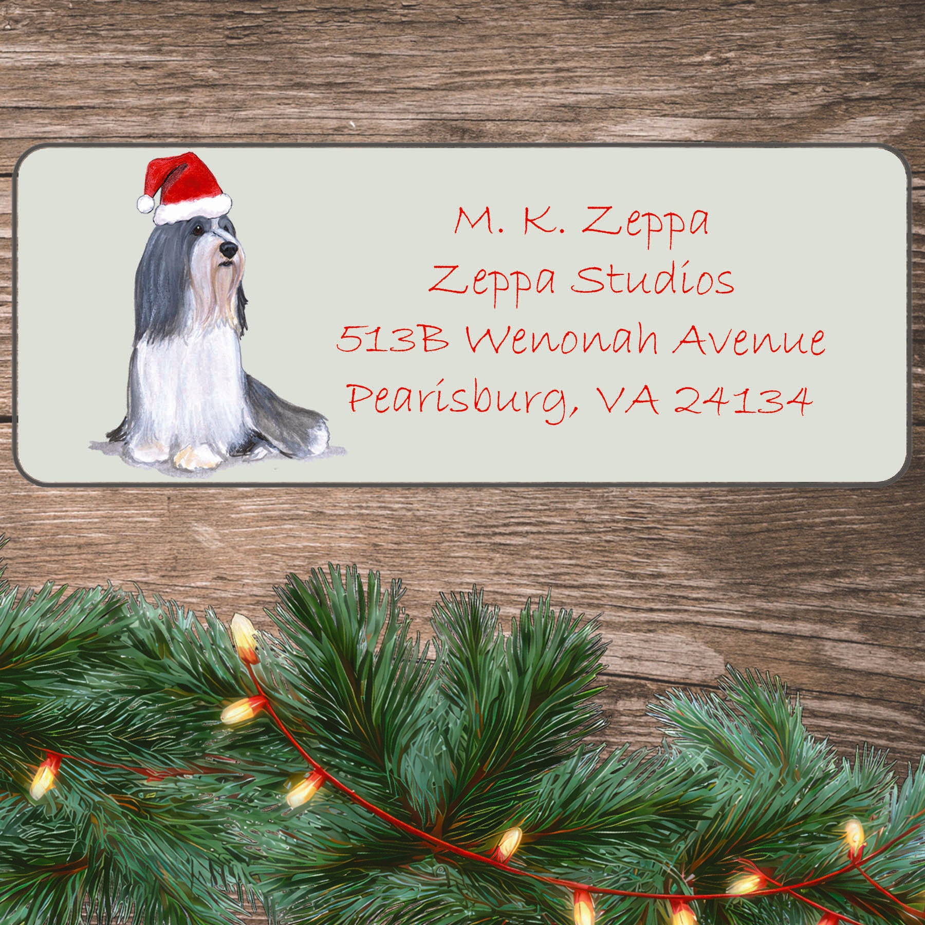 An illustration of a Bearded Collie wearing a Santa Hat on an ivory return address label set off by a wood surface with holiday greenery background.