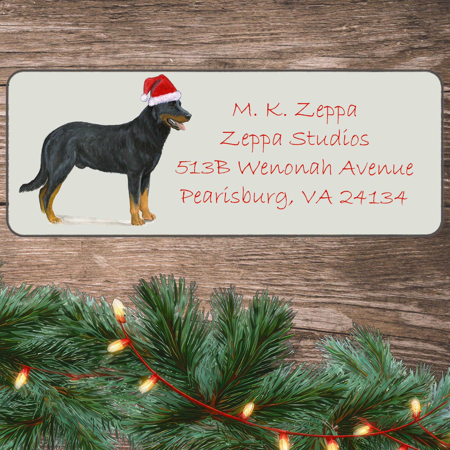 An illustration of a Beauceron wearing a Santa Hat on an ivory return address label set off by a wood surface with holiday greenery background.