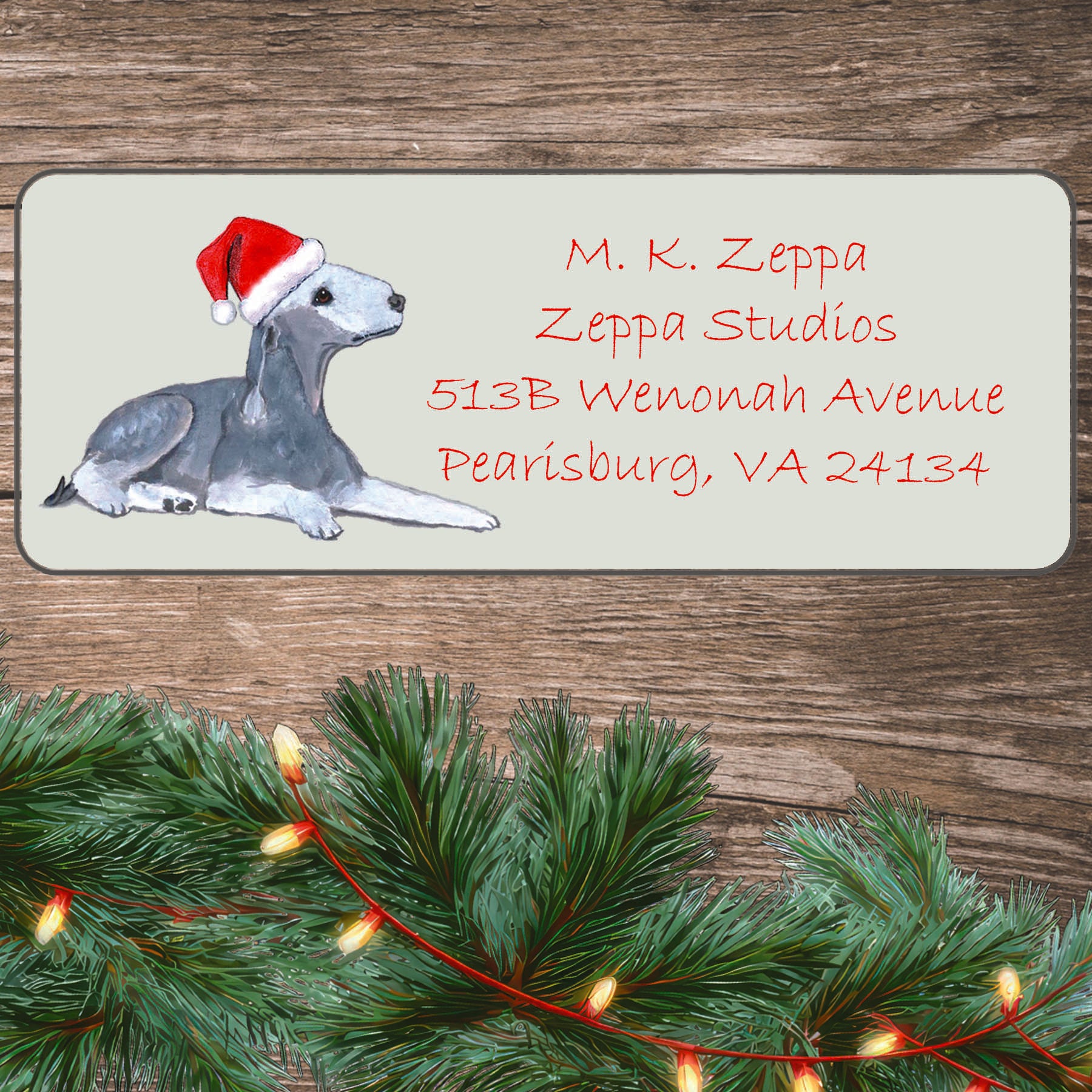 An illustration of a Bedlington Terrier wearing a Santa Hat on an ivory return address label set off by a wood surface with holiday greenery background.