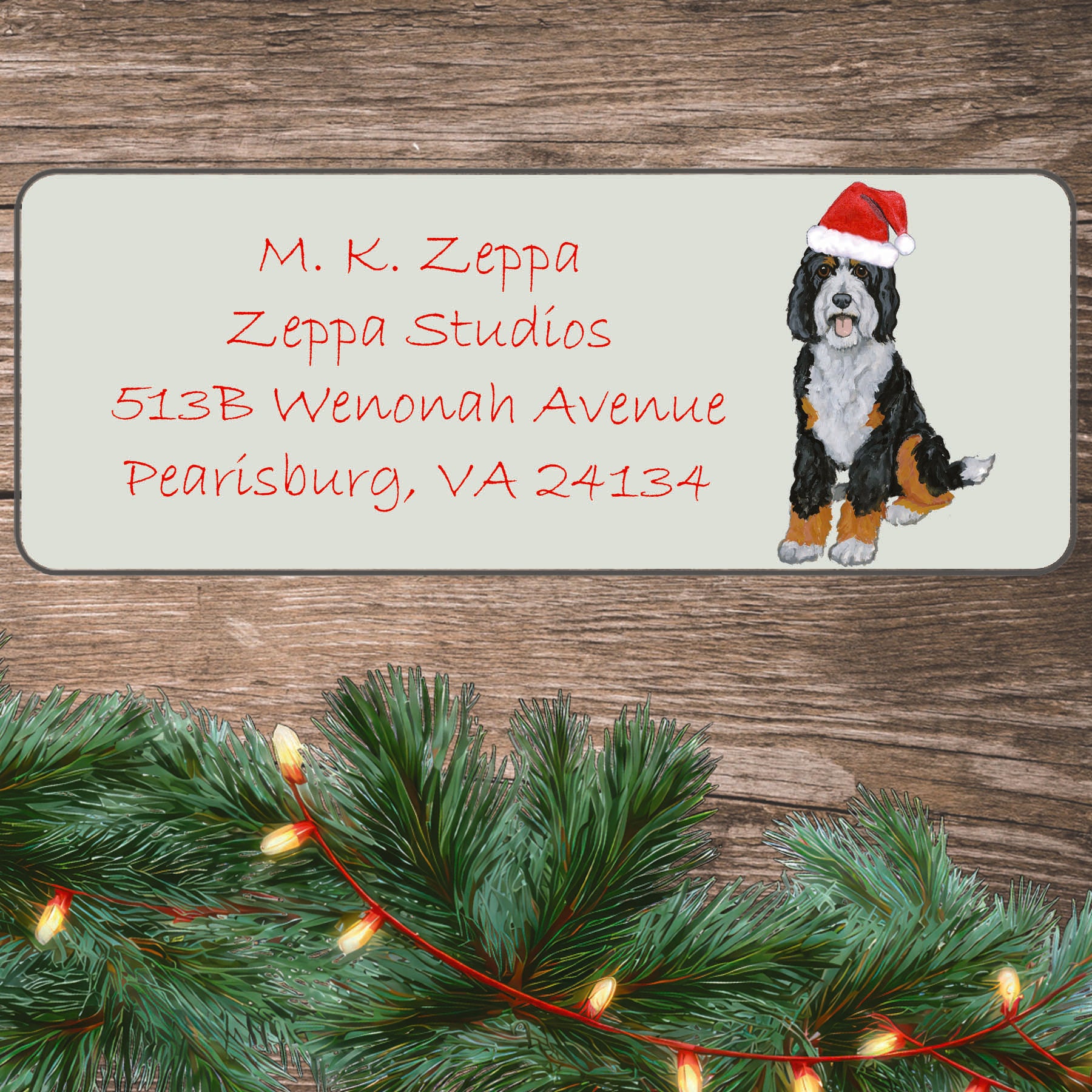 A Bernedoodle wearing a Santa Hat illustration on an ivory return address label set off by a wood surface with holiday greenery background.