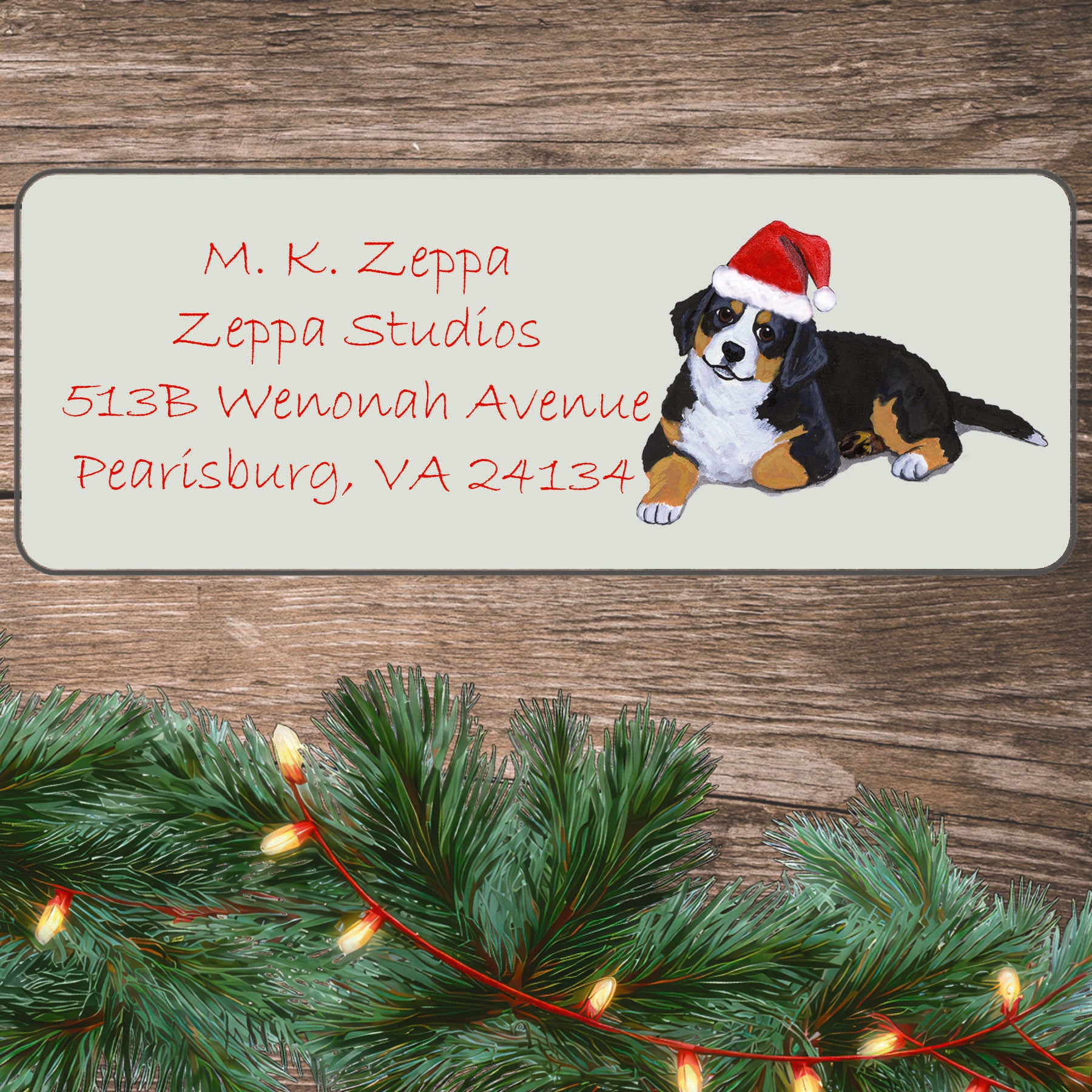 An illustration of a Bernese Mountain Dog puppy wearing a Santa Hat on an ivory return address label set off by a wood surface with holiday greenery background.