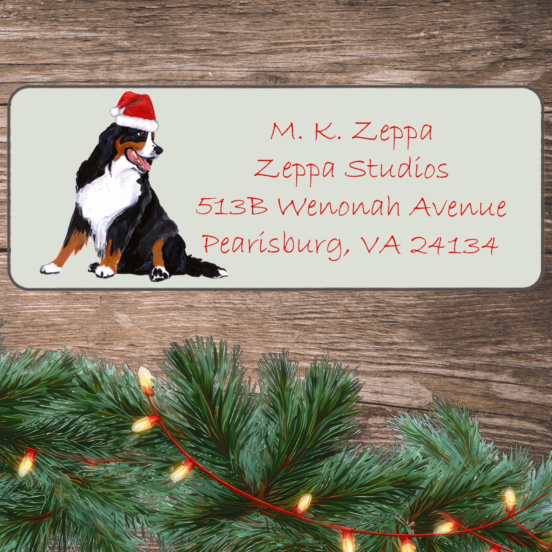 An illustration of a Bernese Mountain Dog wearing a Santa Hat on an ivory return address label set off by a wood surface with holiday greenery background.