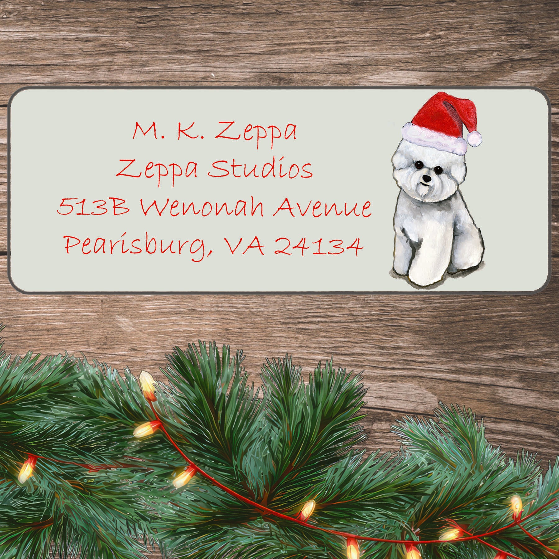 An illustration of a Bichon Frise wearing a Santa Hat on an ivory return address label set off by a wood surface with holiday greenery background.