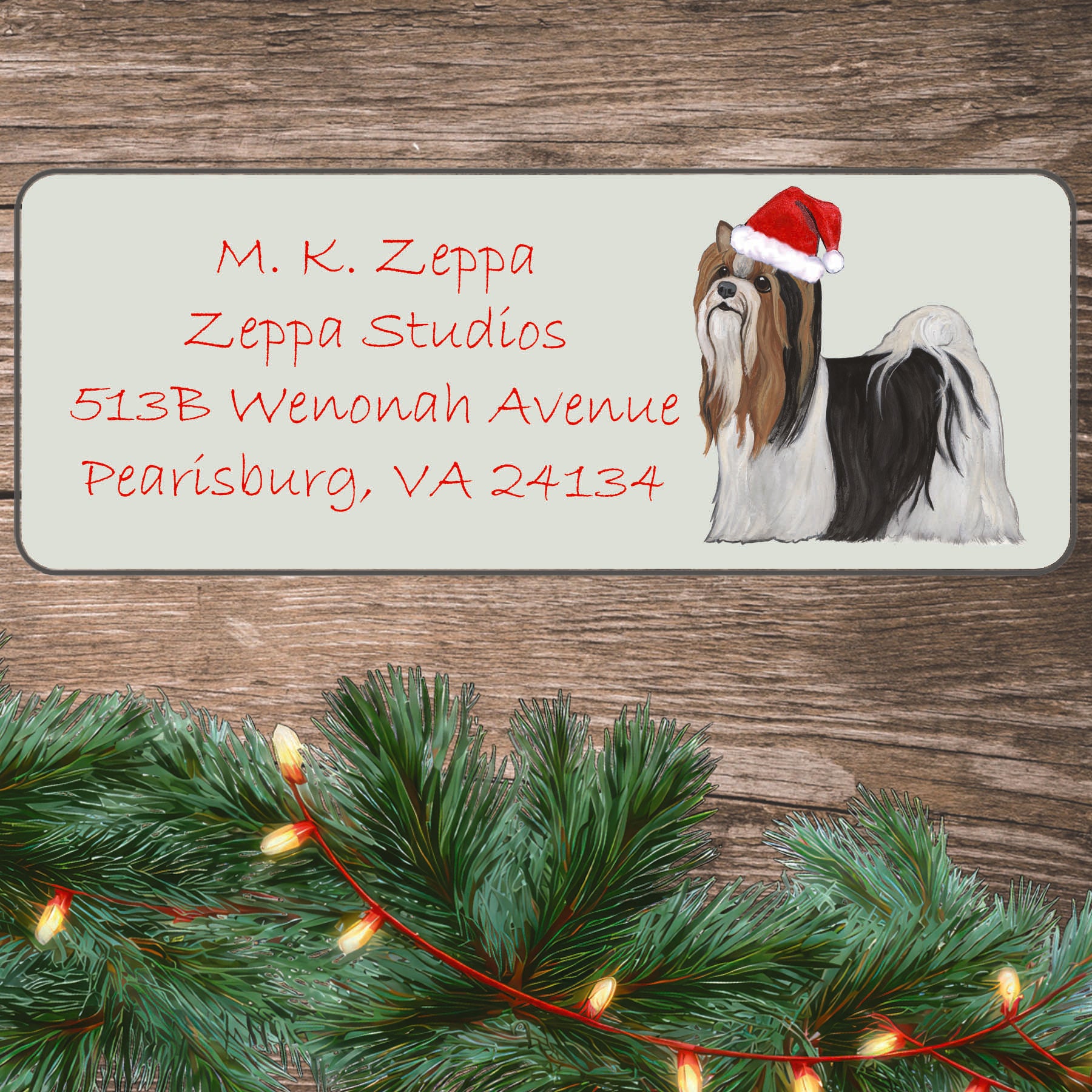 A Biewer Terrier in a Santa Hat illustration on an ivory return address label set off by a wood surface with holiday greenery background.