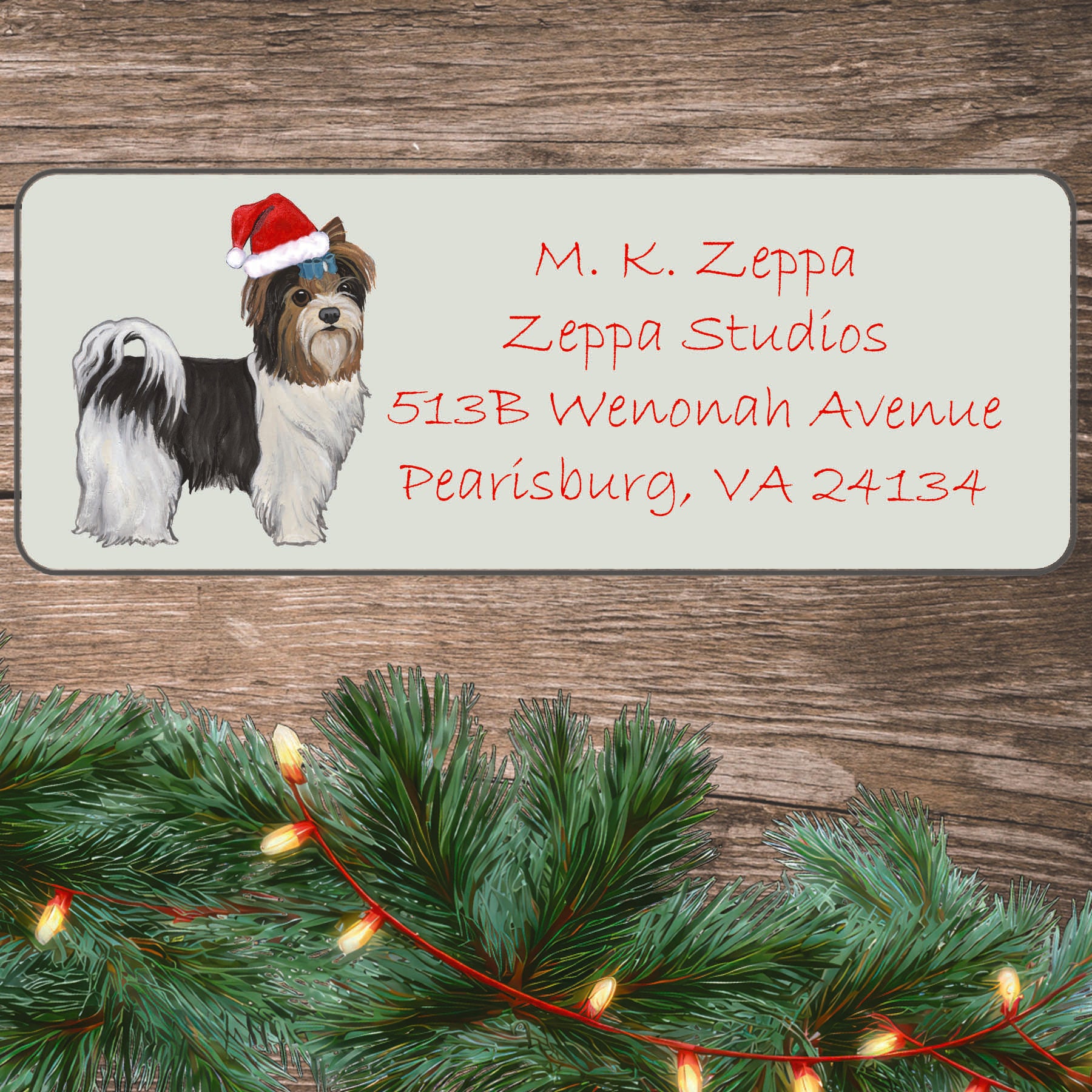 A Biewer Terrier with a short haircut in a Santa Hat illustration on an ivory return address label set off by a wood surface with holiday greenery background.
