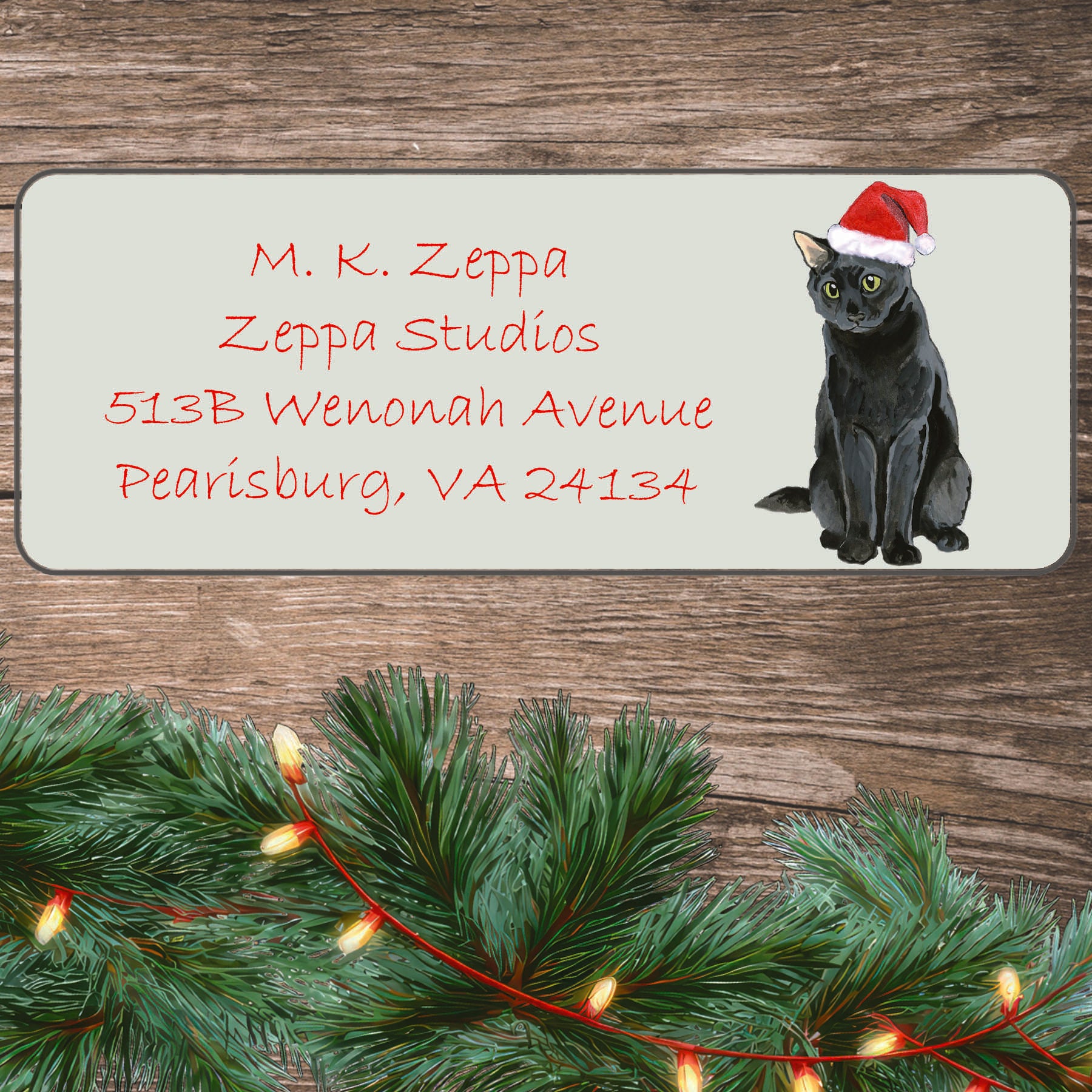 An illustration of a Black Cat wearing a Santa Hat on an ivory return address label set off by a wood surface with holiday greenery background.