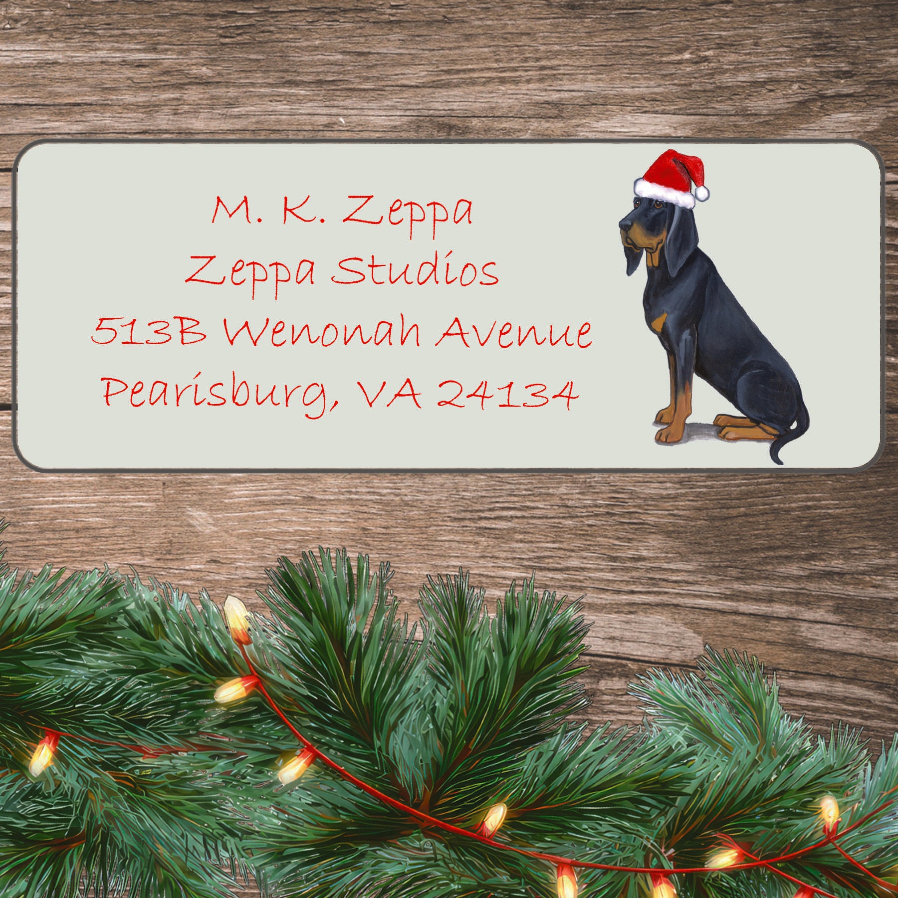 An illustration of a Black and Tan Coonhound wearing a Santa Hat on an ivory return address label set off by a wood surface with holiday greenery background.