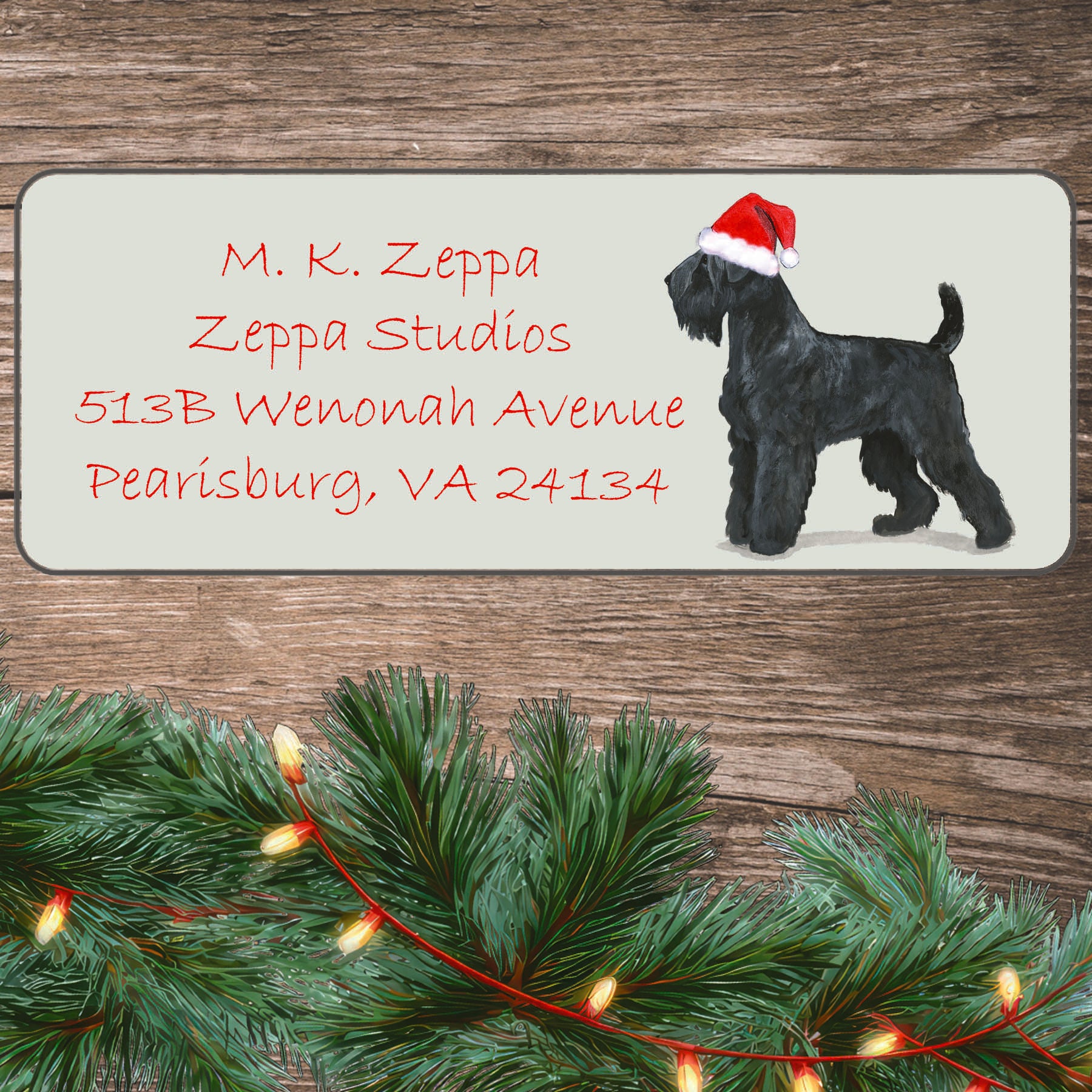 An illustration of a Black Russian Terrier wearing a Santa Hat on an ivory return address label set off by a wood surface with holiday greenery background.