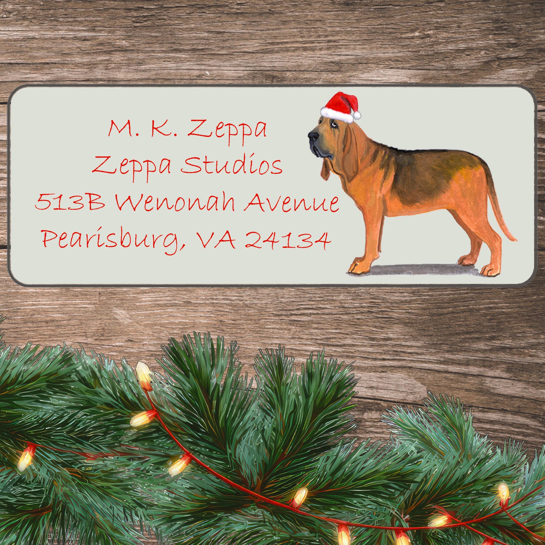 An illustration of a Bloodhound wearing a Santa Hat on an ivory return address label set off by a wood surface with holiday greenery background.