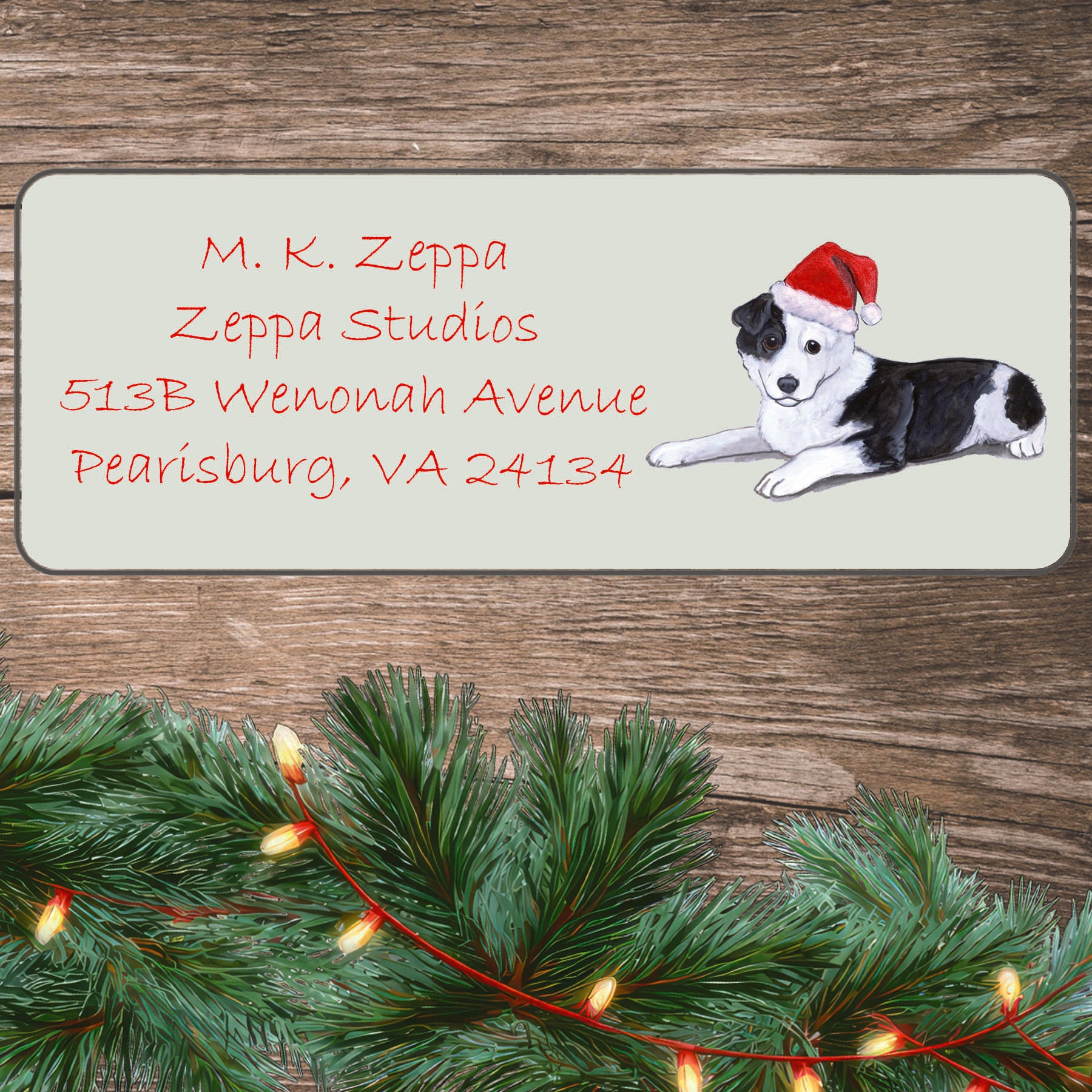 An illustration of a Border Collie puppy wearing a Santa Hat on an ivory return address label set off by a wood surface with holiday greenery background.